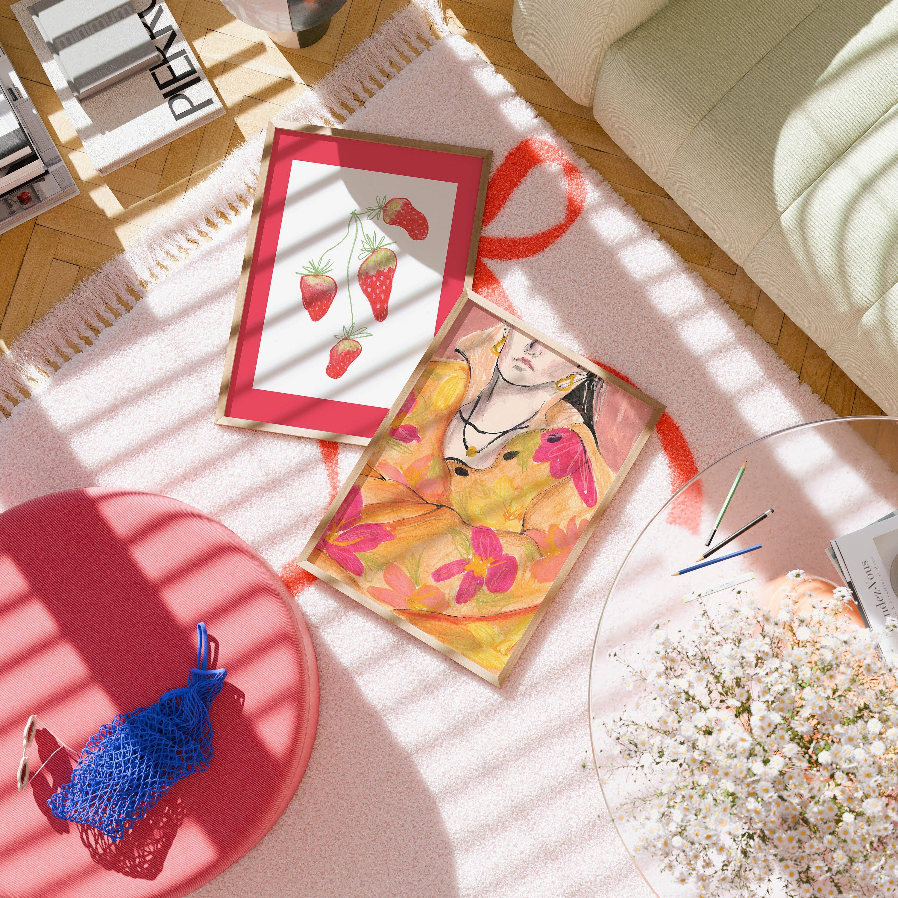 Two framed art prints displayed on a light pink rug in a sunlit room. One print features red strawberries on a white background with a red frame, and the other features a colourful portrait with yellow, pink, and orange tones.