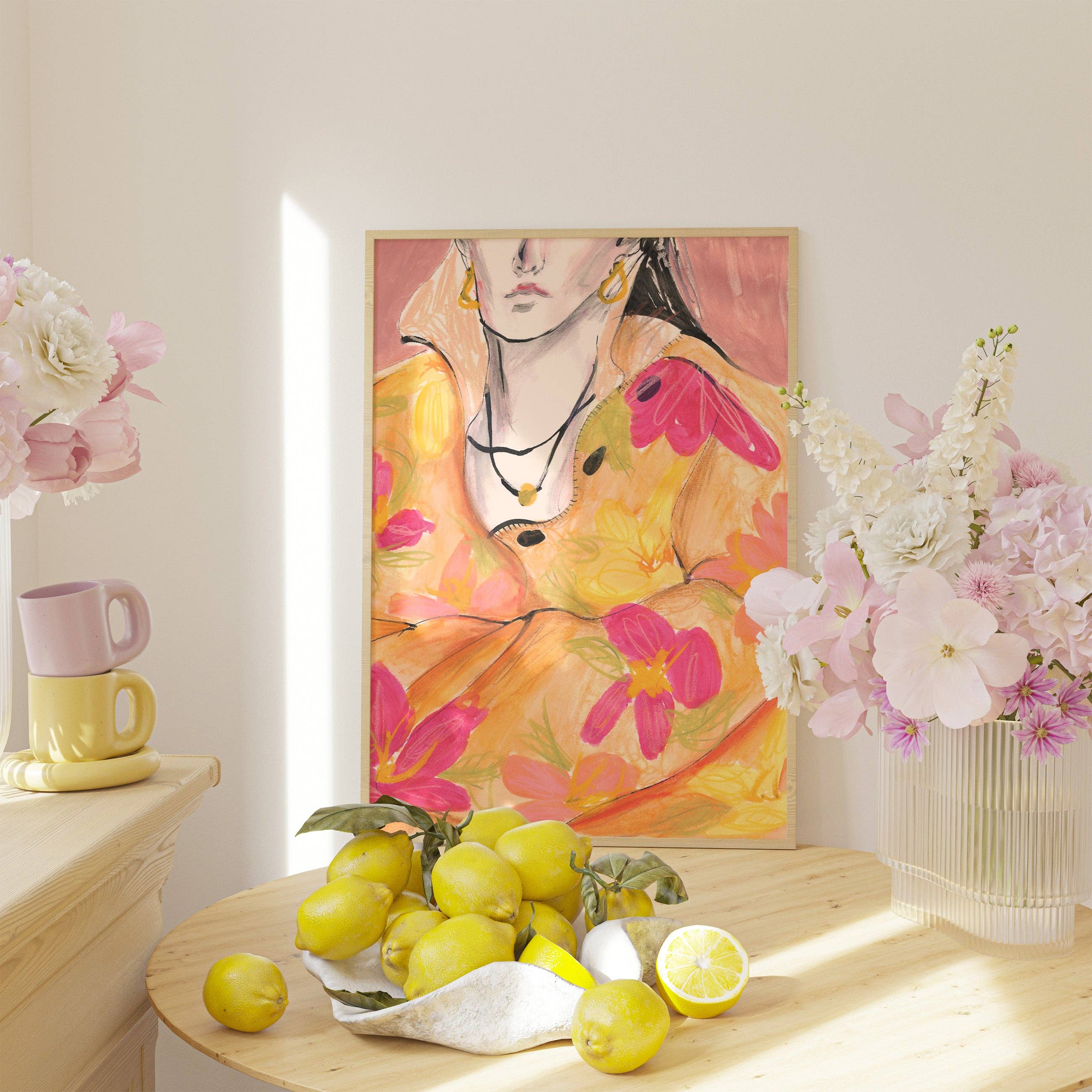 A framed art print featuring a colorful painting of a woman in a yellow dress with pink flowers, styled on a light wood table with lemons and flanked by pink and white flowers in a sunlit room.