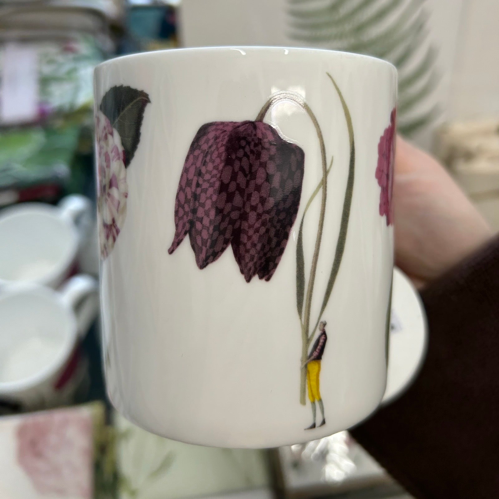White mug with purple flower design held by a hand in a blurred indoor setting.