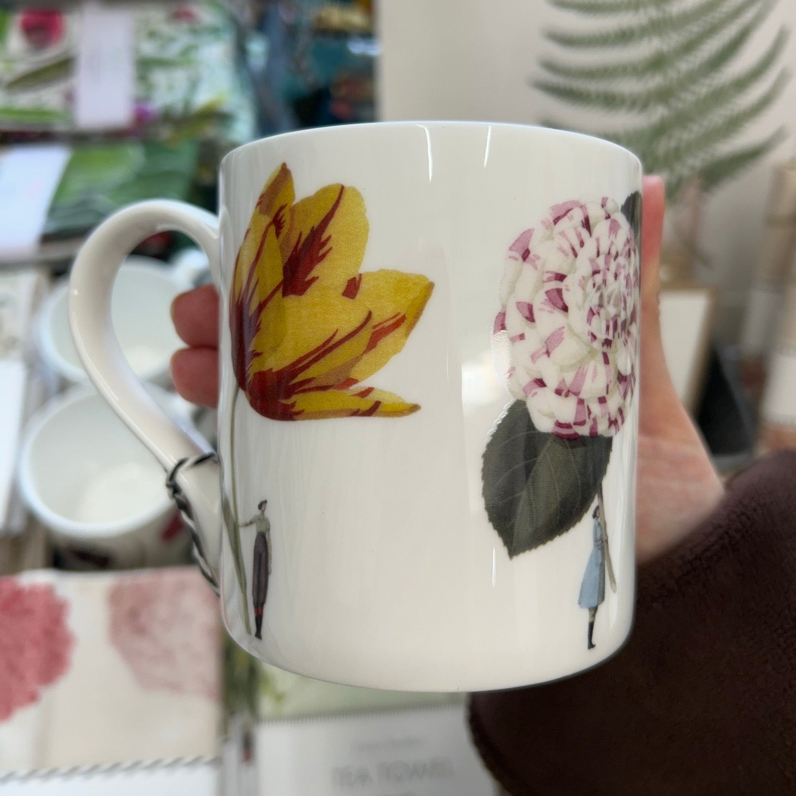 White mug with floral designs held by a hand, on a blurred background.