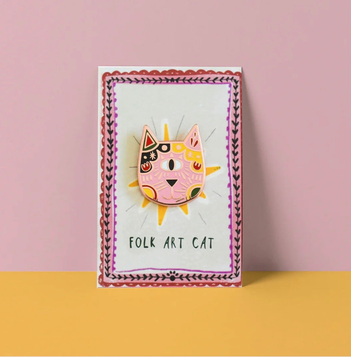 Folk art cat pin on a card with decorative border against a pink background.