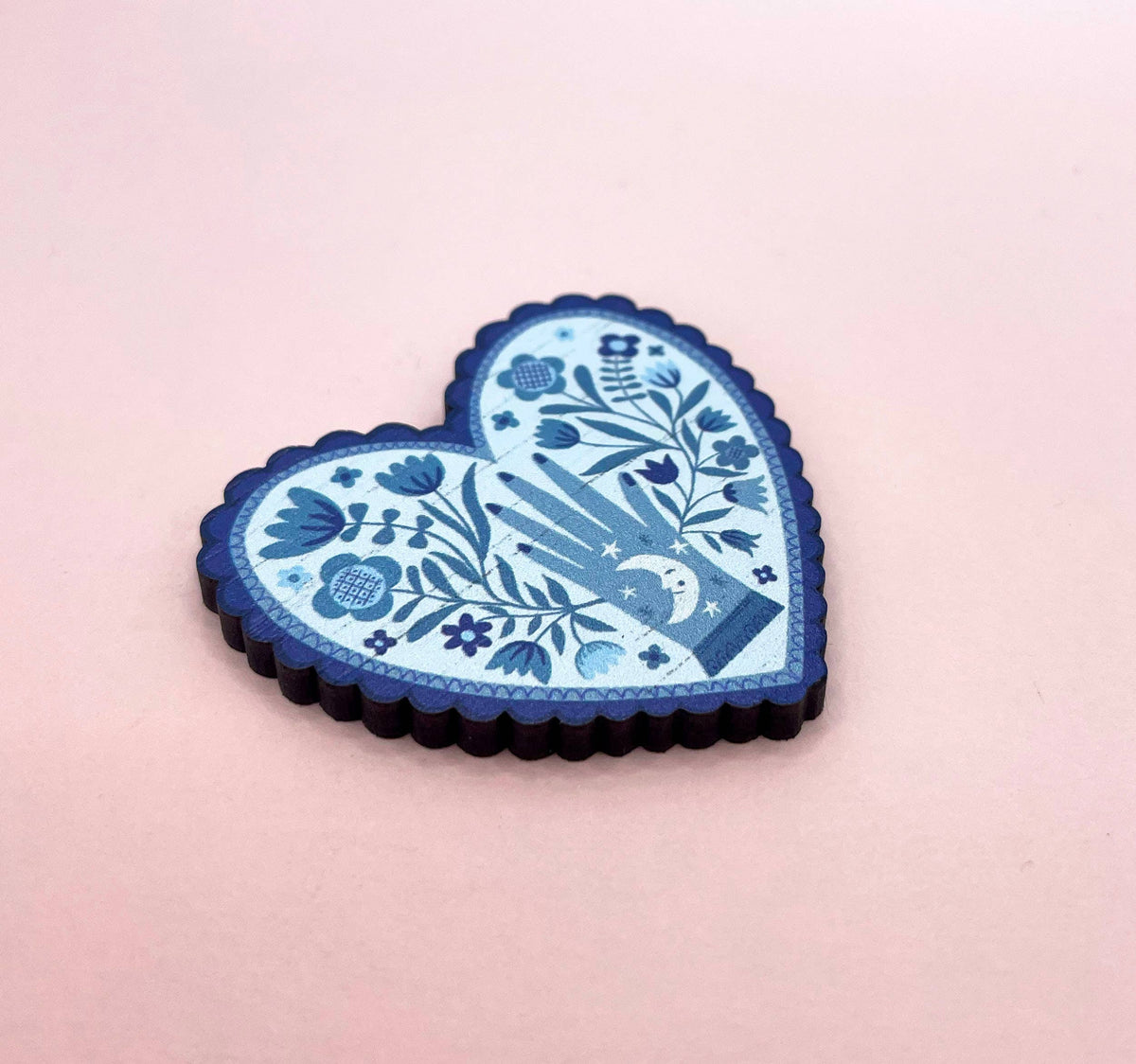 Decorative heart-shaped blue brooch with floral and hand design on a pink background.