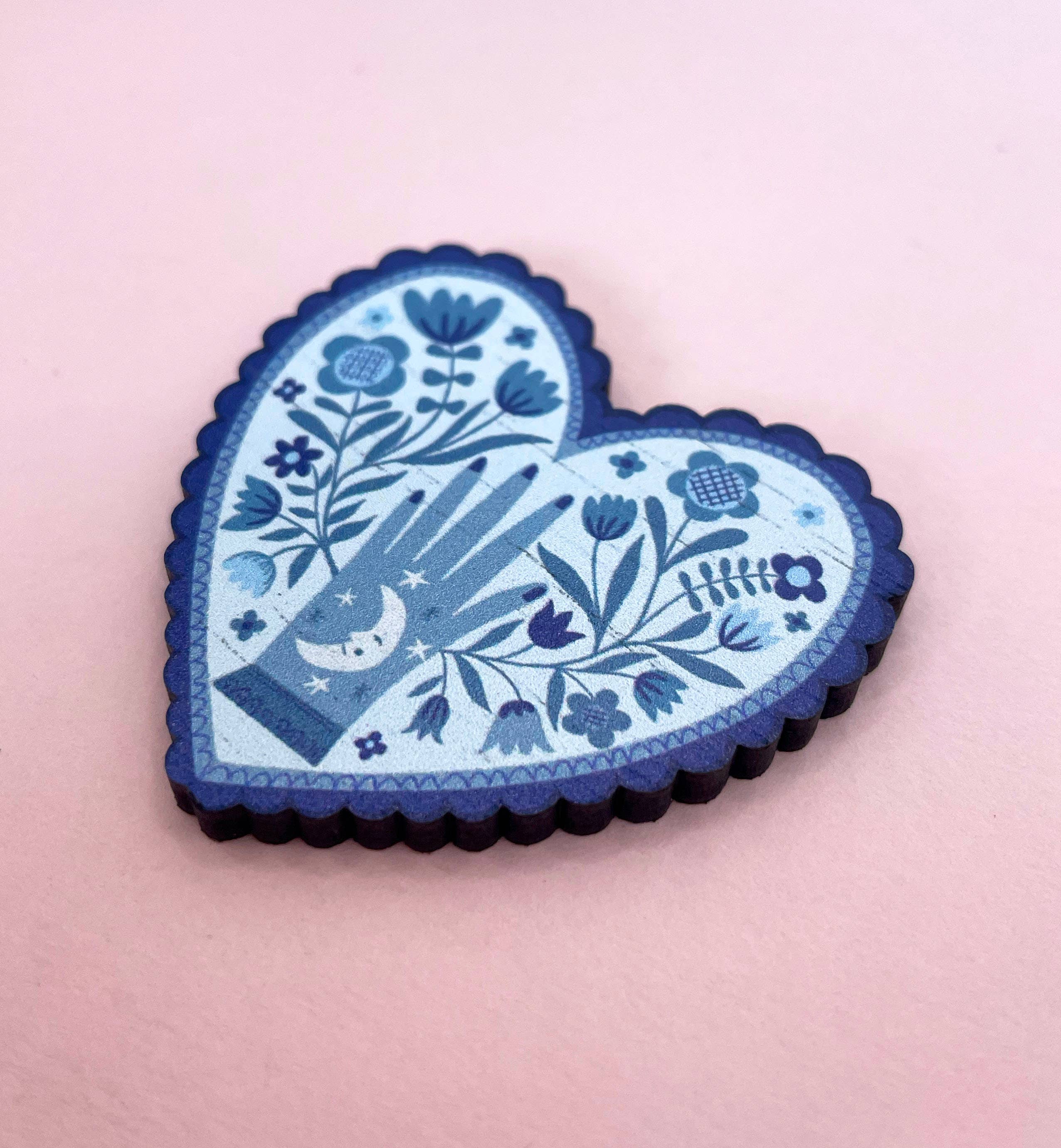 Heart-shaped blue brooch with floral and hand design on a pink background.