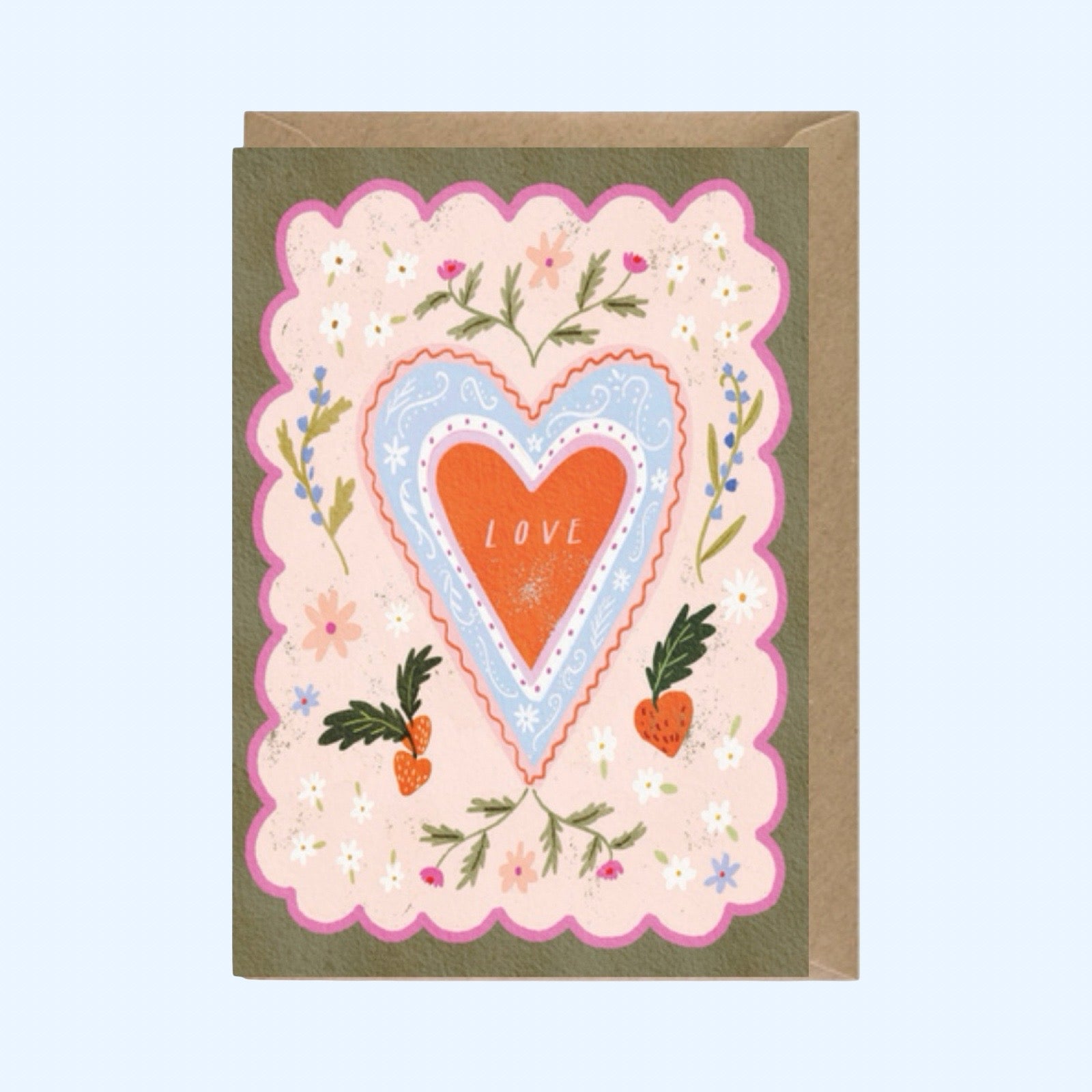 A greeting card with a brown envelope behind it, featuring a decorative floral heart design. The central heart is orange with the word 'LOVE' in white, surrounded by a light blue scalloped heart and a larger pink scalloped border with small flowers and green leaves, all on a pale blue background.