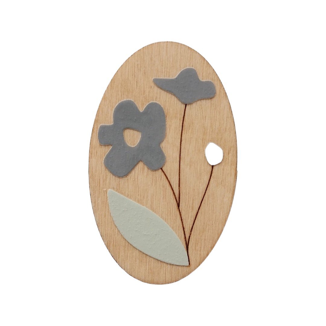 An oval wooden brooch with a blue forget me not flower, brown stem and green leaf.