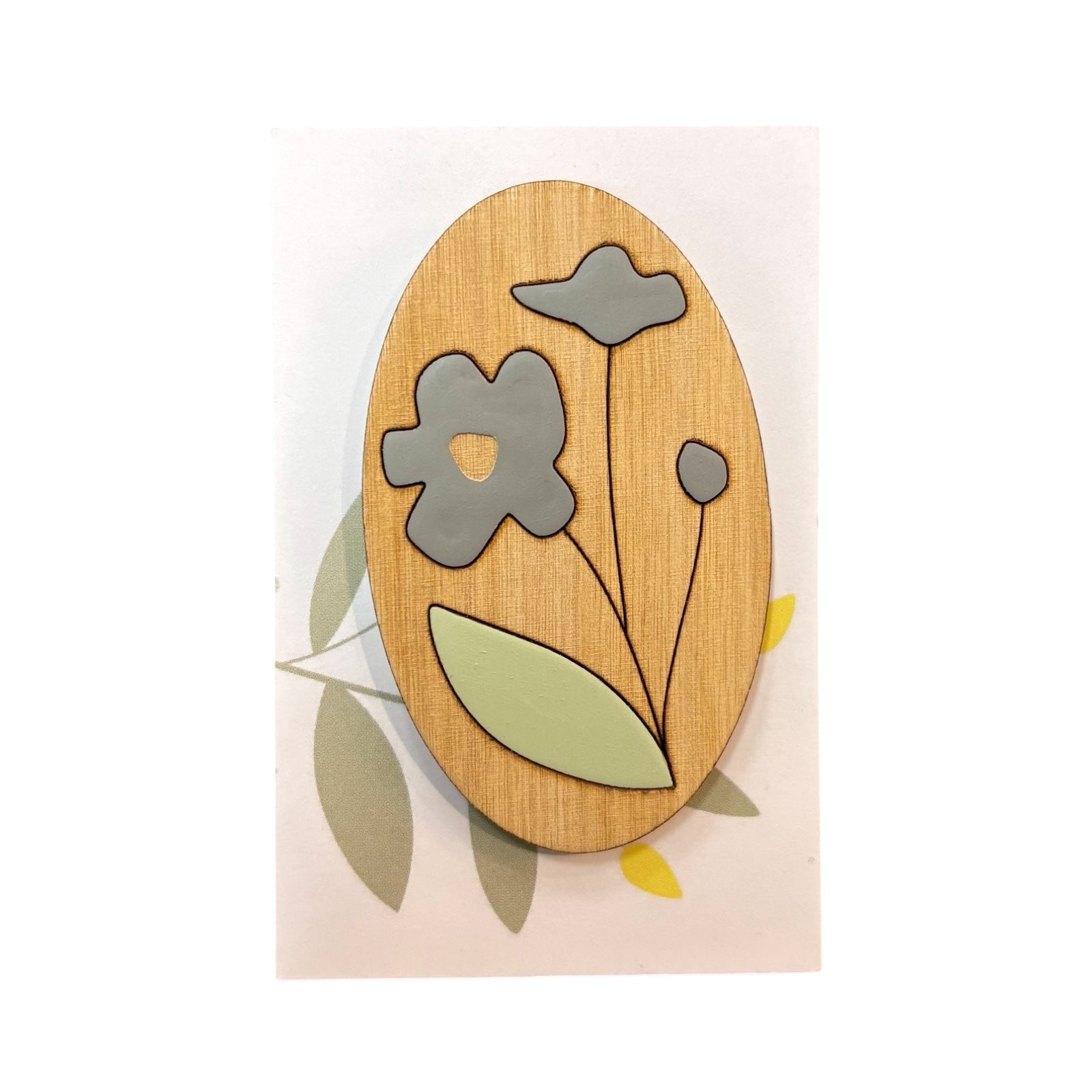 An oval wooden brooch with a blue forget me not flower, brown stem and green leaf on a white backing card.