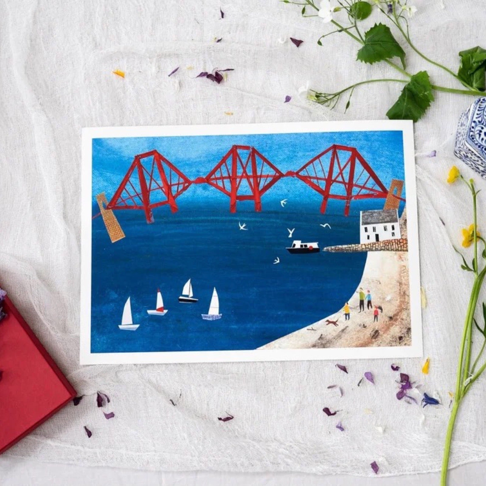 Illustration of a red bridge over water with sailboats and a beach on a fabric white background surrounded by petals and a red notebook.