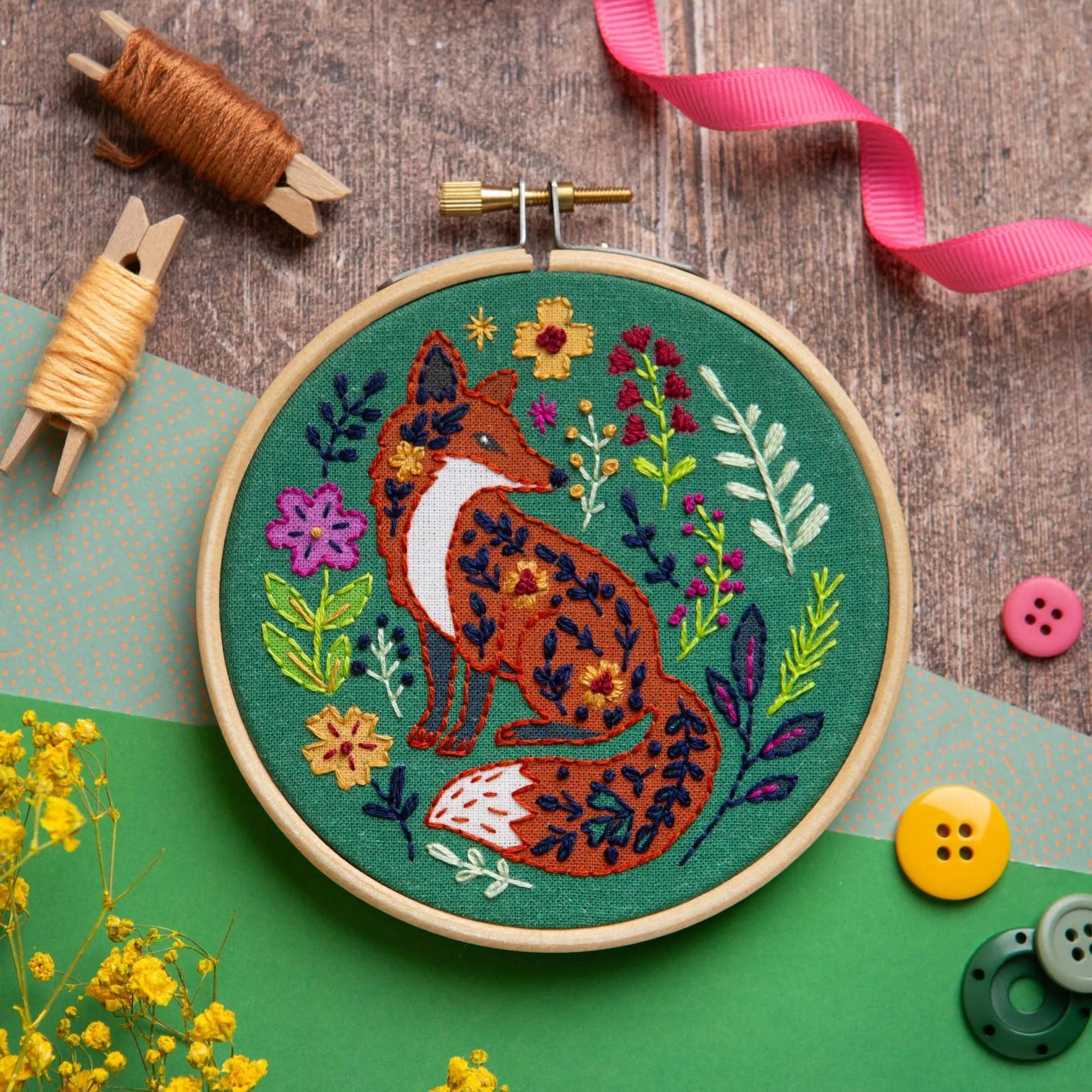 A circular embroidery hoop displaying a finished embroidered design of a fox surrounded by flowers on a green background, styled with spools of thread, buttons, and ribbon on a wood and green surface.