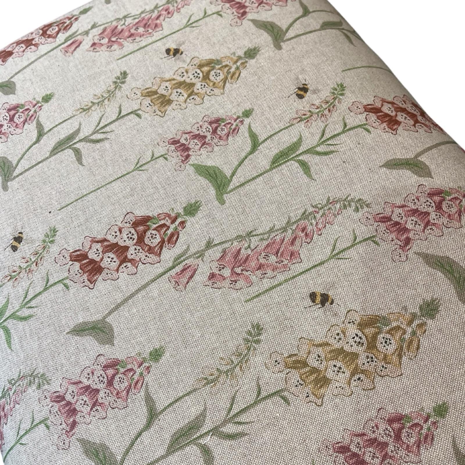 A rectangular bench with cream linen fabric upholstery featuring a floral pattern of foxgloves in pink, red, and yellow, with honey bees. It has mahogany legs and is finished with matching binding.