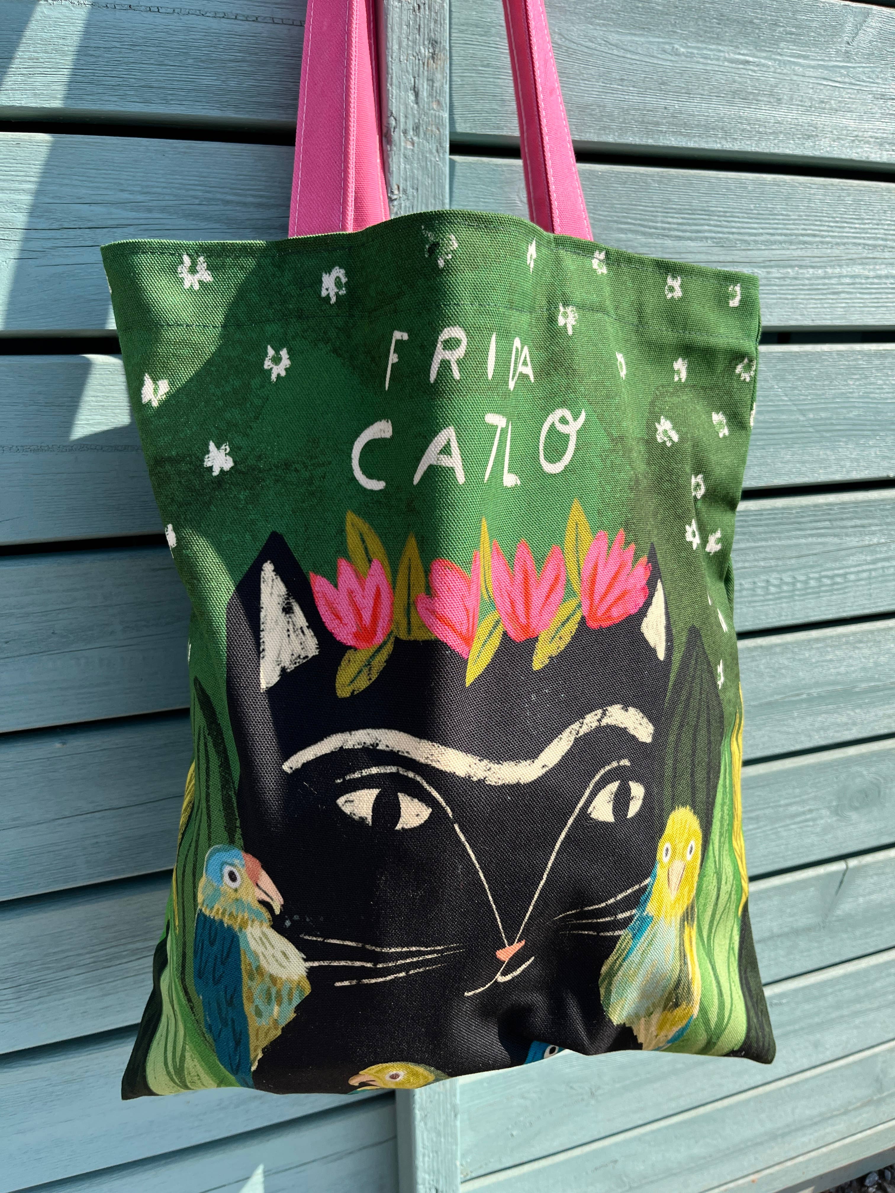 A green canvas tote bag with pink handles, featuring a black cat face, pink flowers, birds, and the text 'FRIDA CATLO' on a blue wooden slat background.
