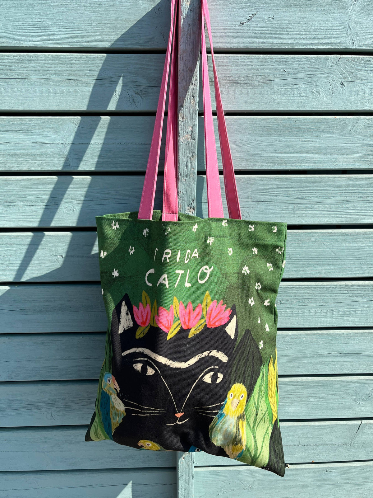 A tote bag with pink handles hanging against a light blue wooden wall. The bag features a green background with white text reading 'FRIDA CATLO', a large black cat face, pink and white flowers.