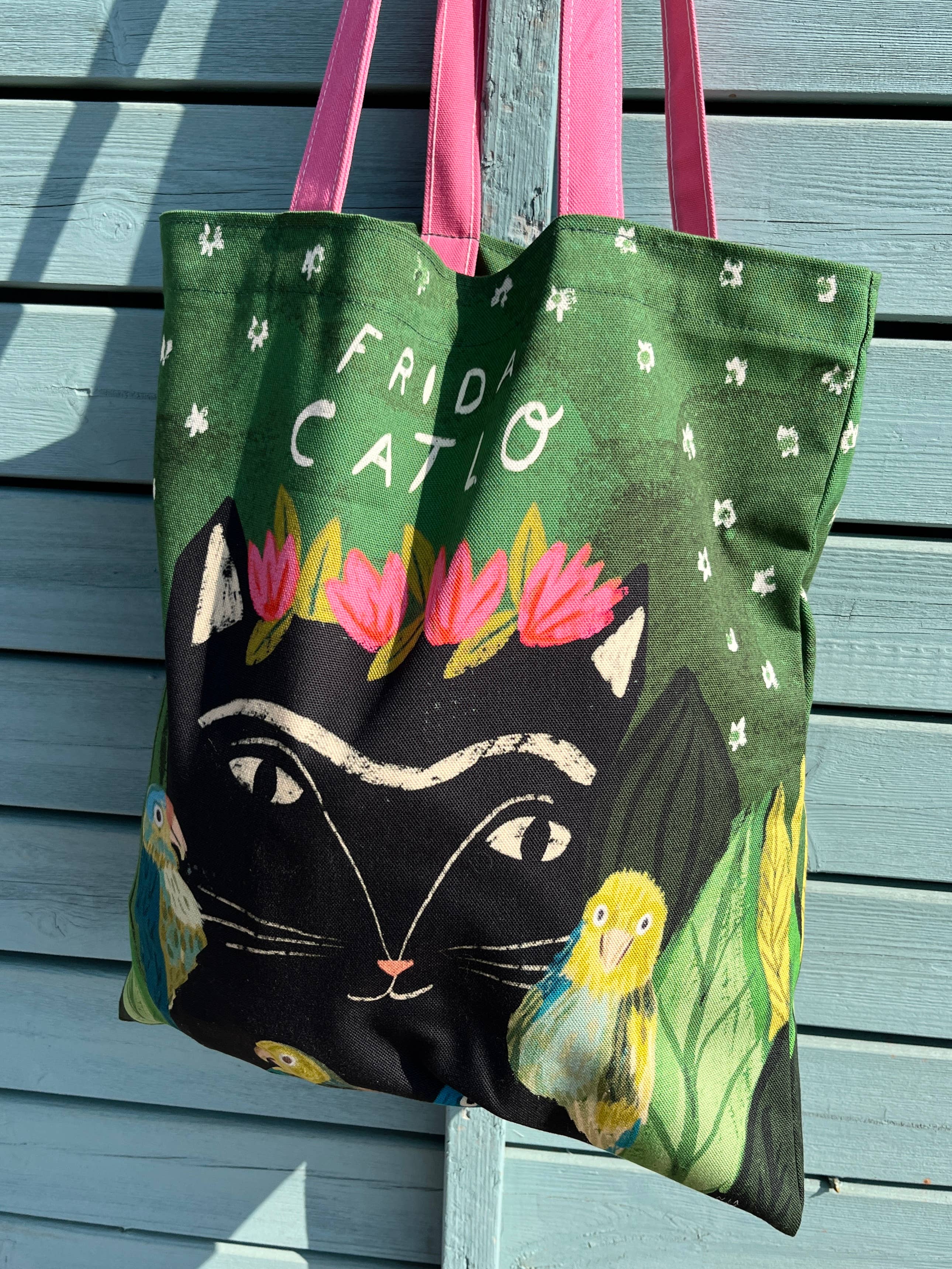 A green canvas tote bag with pink handles, featuring an illustration of a black cat wearing a pink flower crown, with the words 'FRIDA CATLO' printed above. The bag is shown hanging against a light blue wooden slat wall.