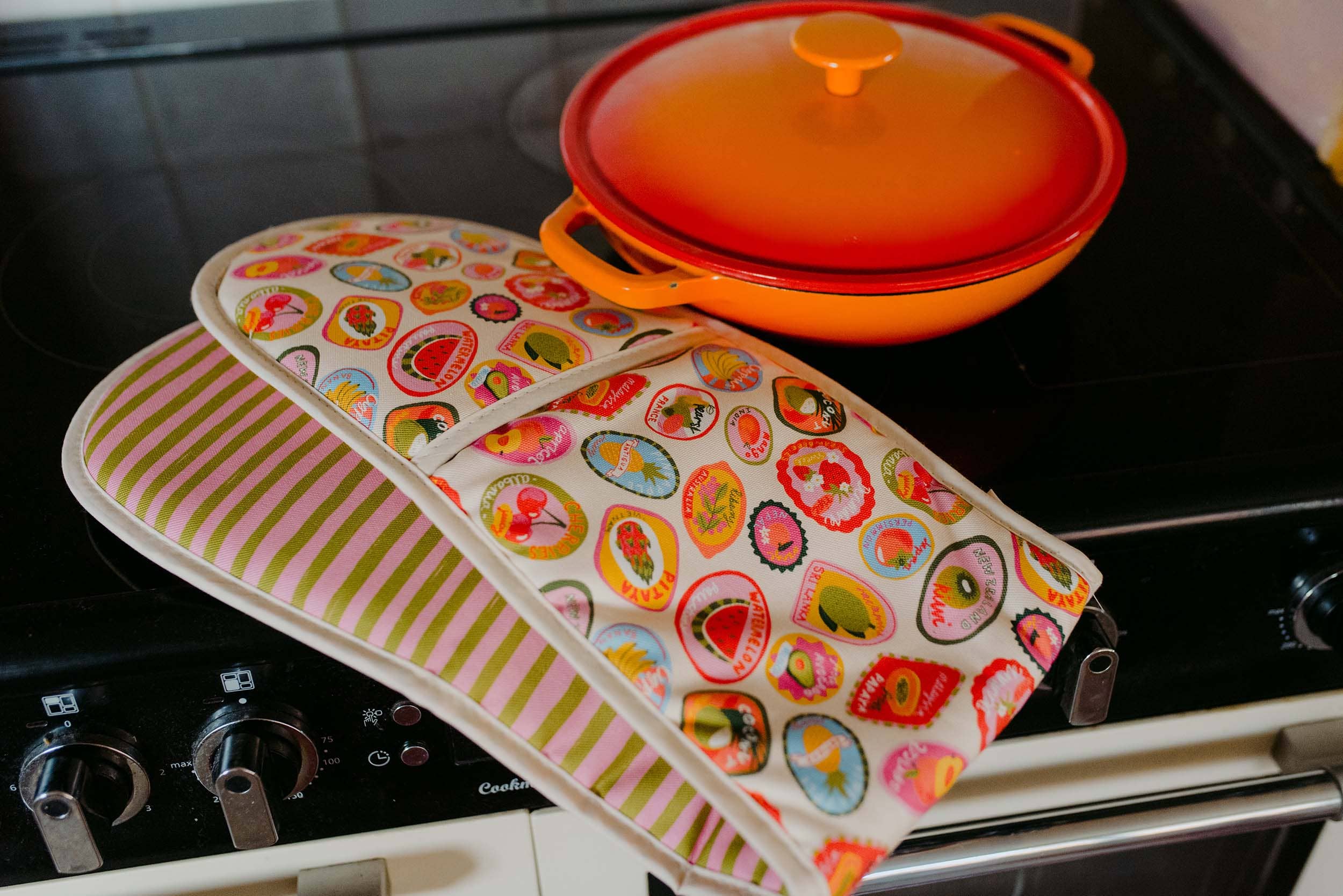 Colorful oven mitt and pot holder on a stovetop with a lid in the background.