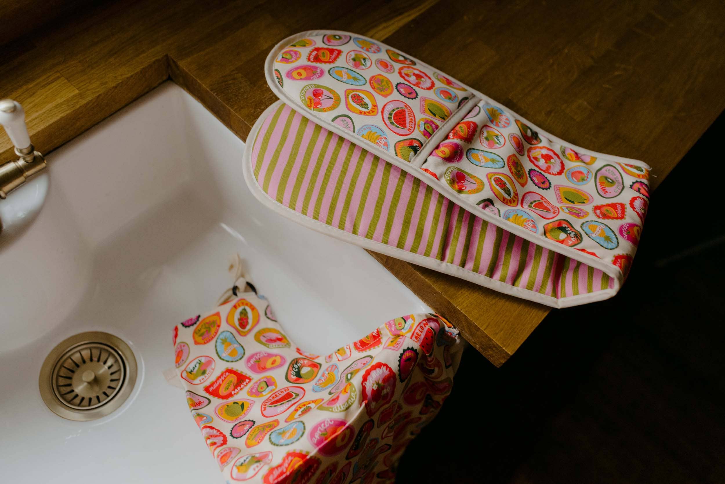 Colorful oven mitts with fruit pattern on a kitchen counter next to a sink.