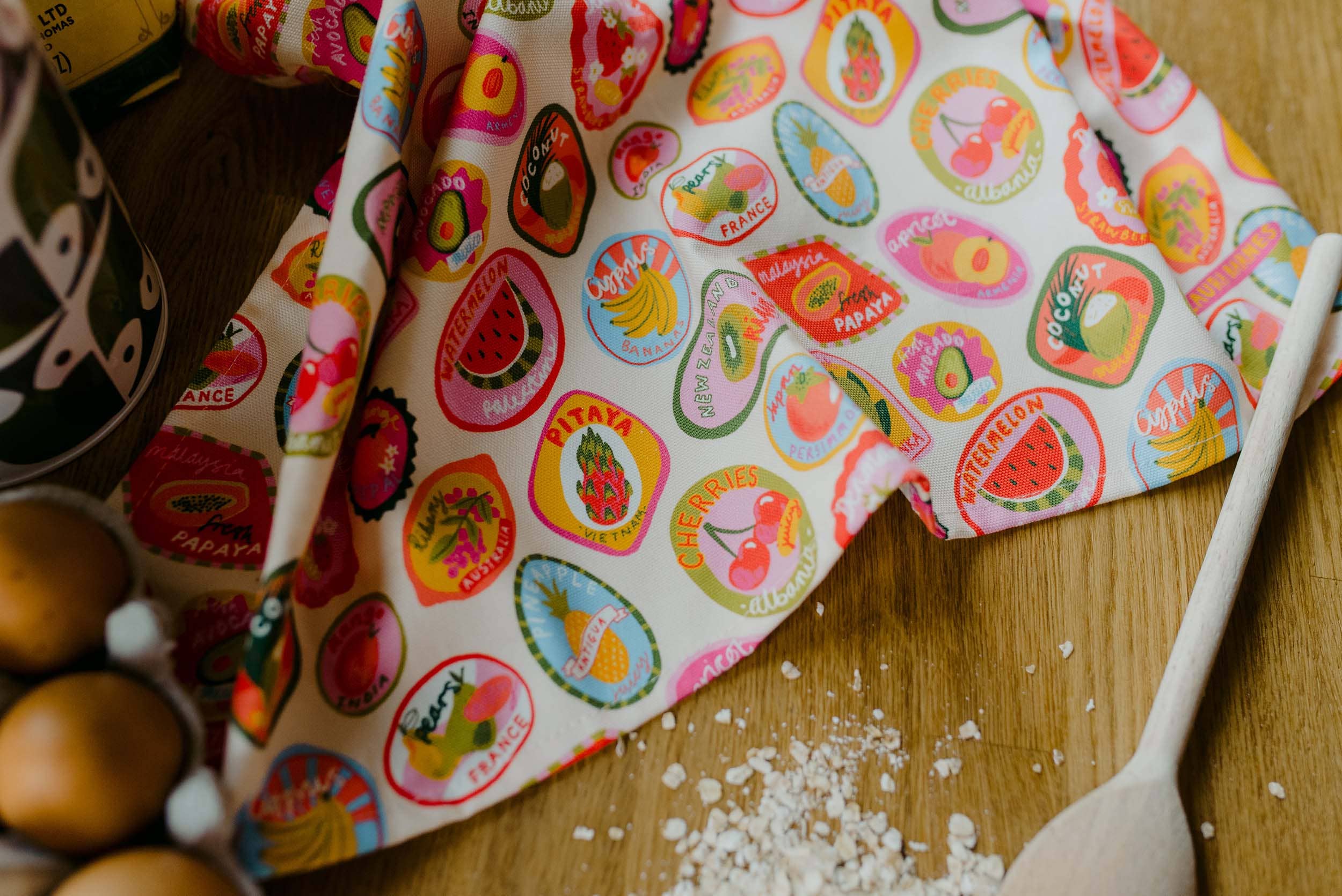 Colorful kitchen towel with fruit pattern on a wooden surface with eggs and a spoon.