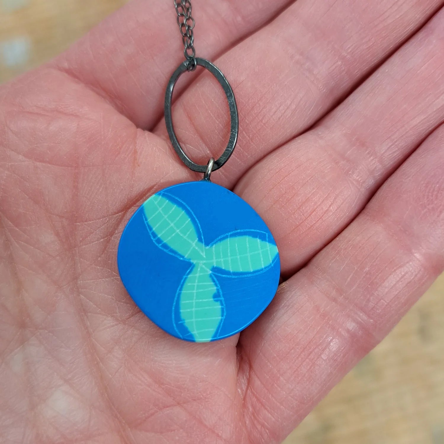 Blue circular pendant with a green leaf design held in a hand.