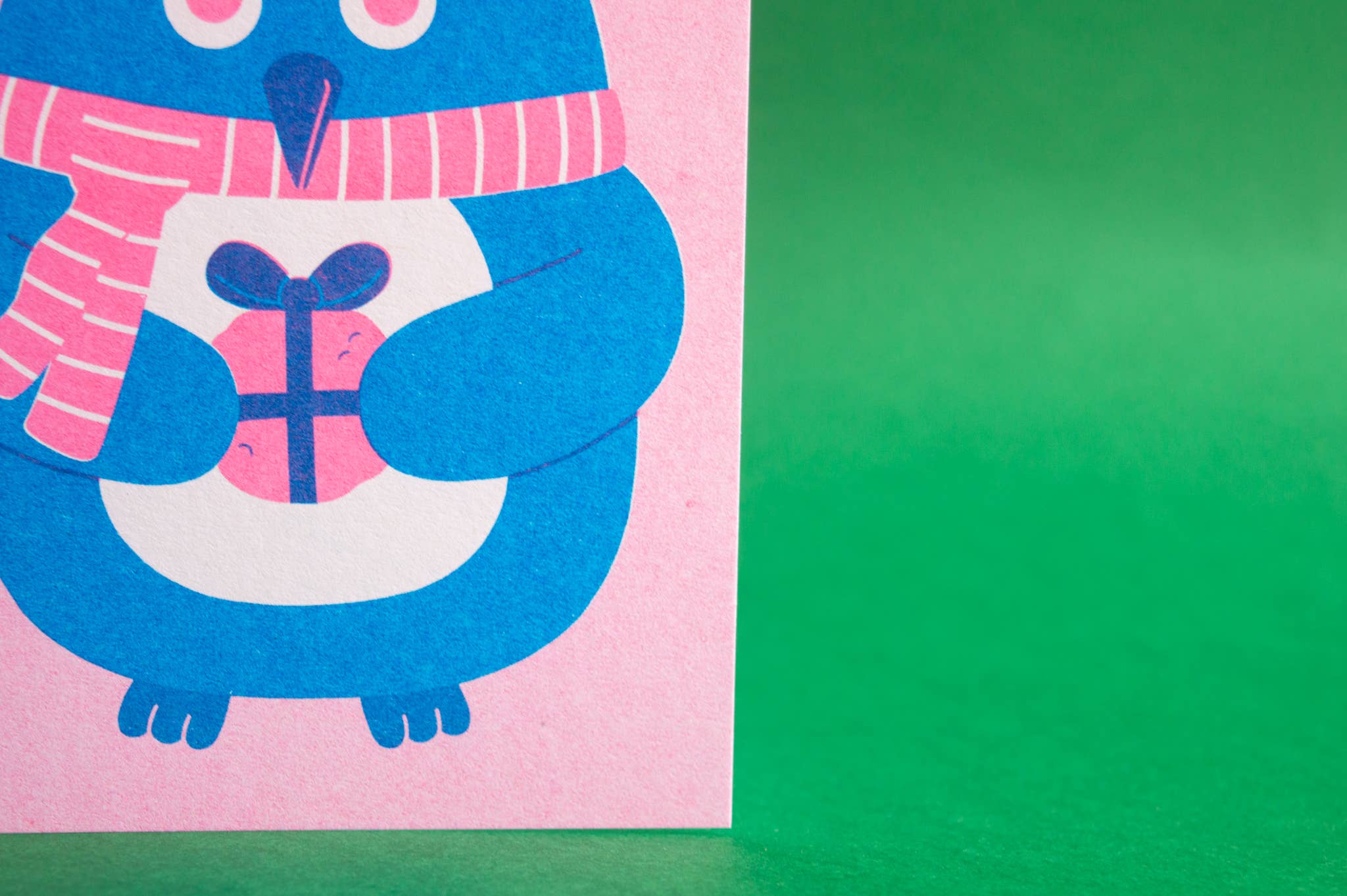 A close-up of a pink card featuring a blue penguin wearing a pink striped scarf and holding a pink gift box with a dark purple bow. The card is positioned on a solid green surface.