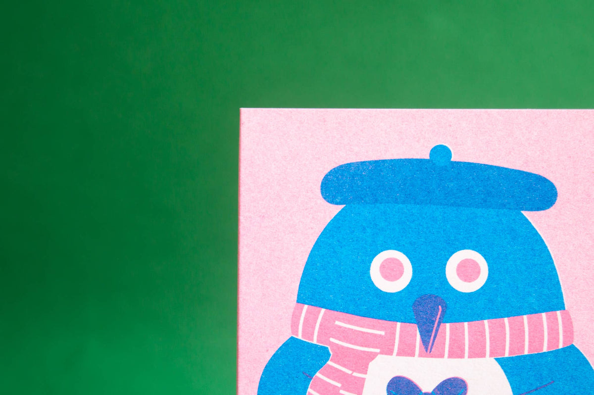 A close-up of a colourful art print featuring a blue penguin wearing a pink striped scarf and a blue beret, set against a pink card on a green background.