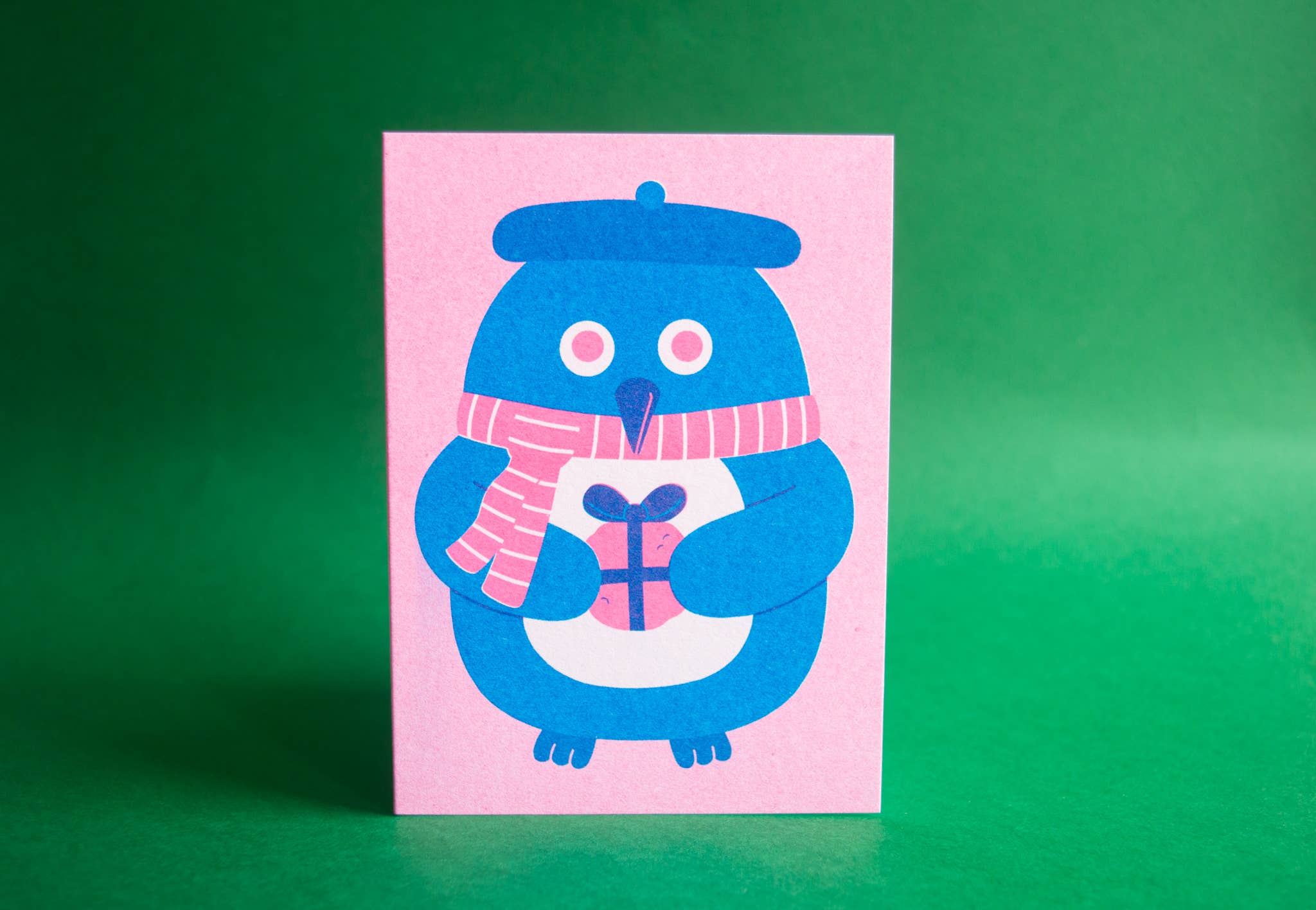 A square pink greeting card featuring a stylized blue penguin wearing a pink striped scarf and holding a wrapped gift, against a solid green background.