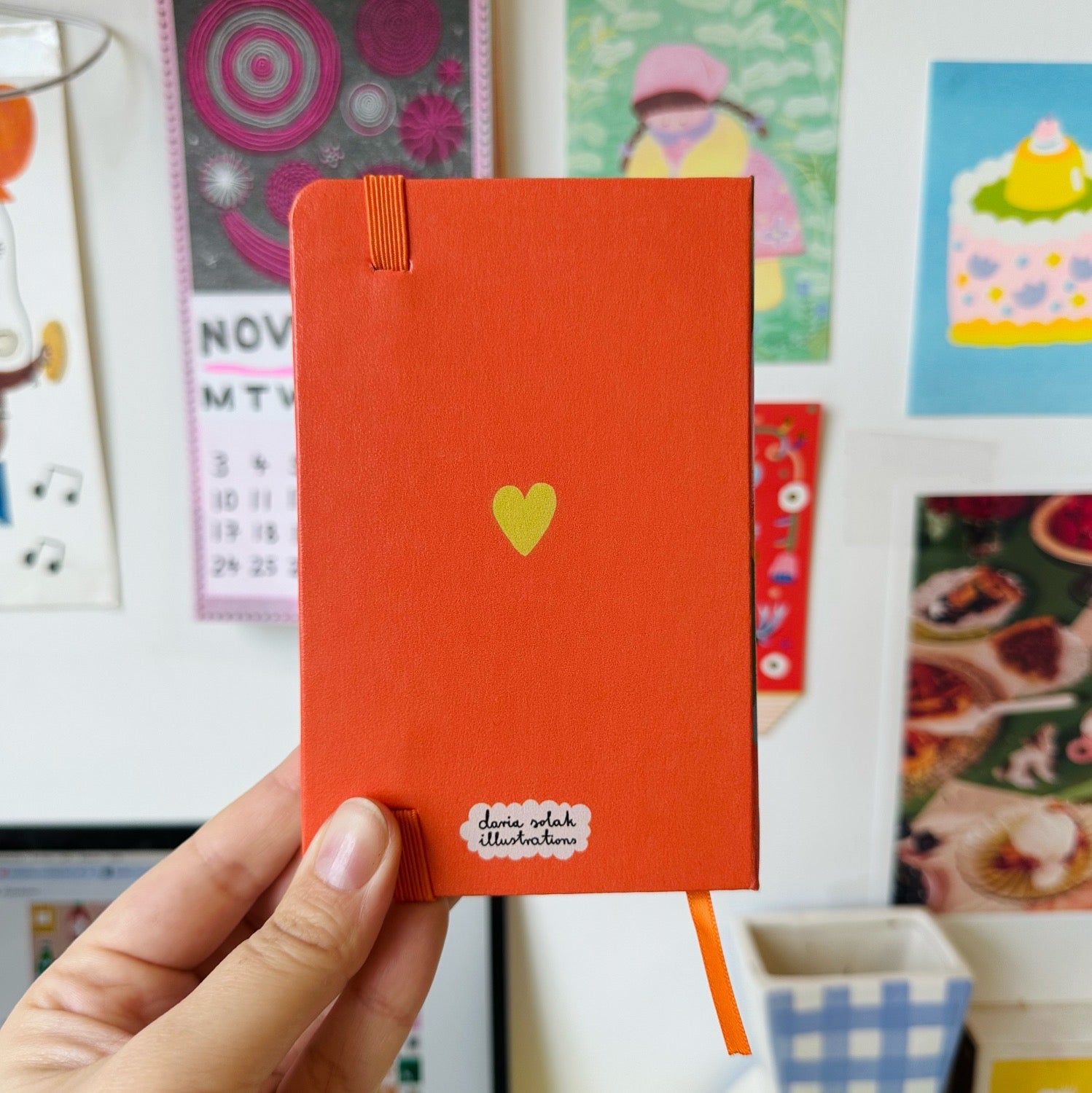 Red notebook with a yellow heart held in front of a wall with various colorful posters.