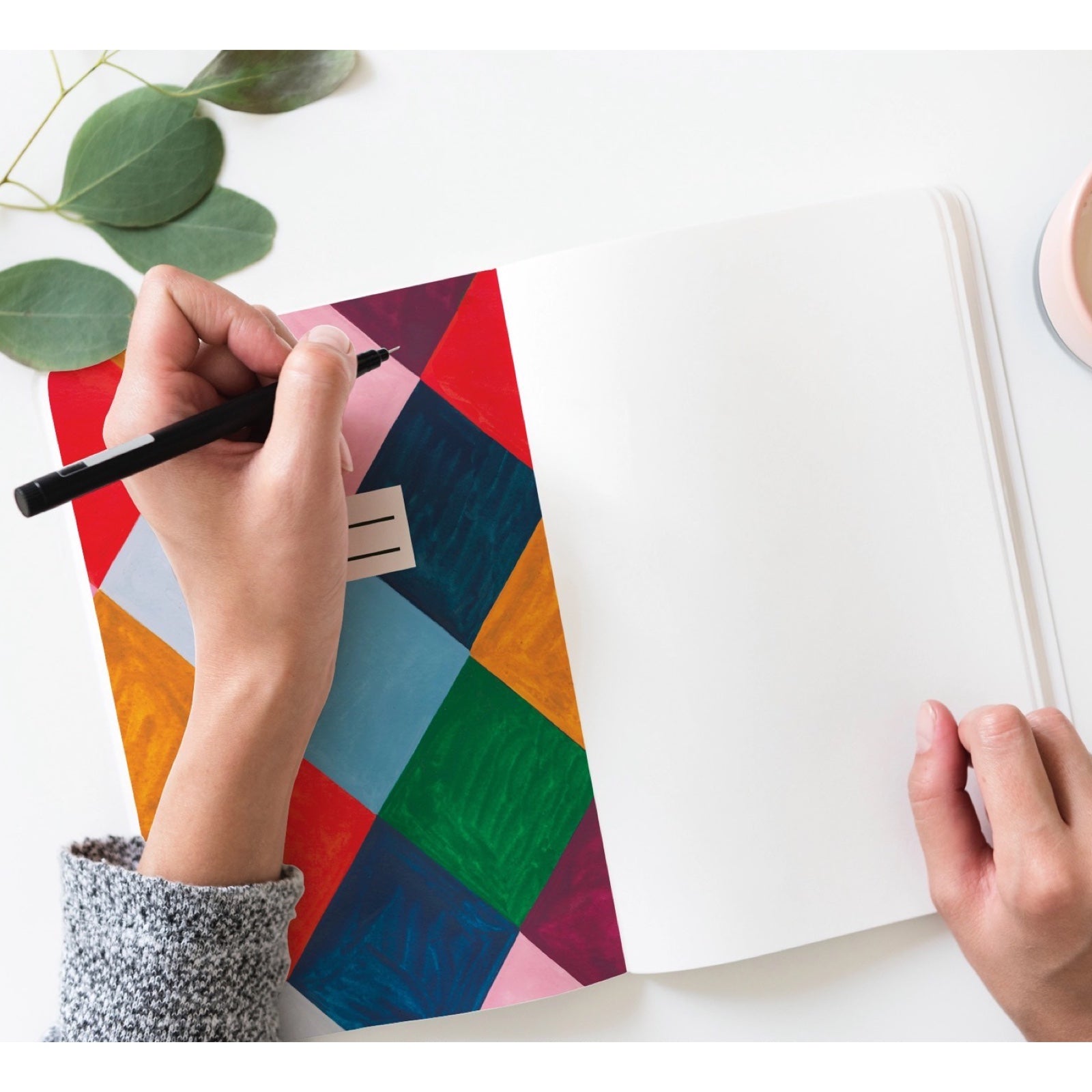 Hand holding a pen over a colorful geometric-patterned paper on a white surface with green leaves.