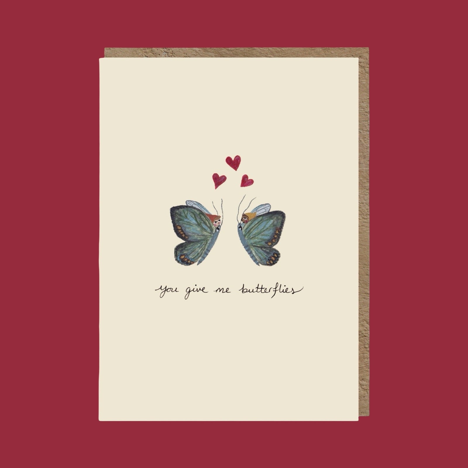 A cream-colored greeting card with a brown envelope, featuring an illustration of two blue butterflies facing each other with three small red hearts above and the handwritten text 'you give me butterflies' below, set against a solid dark red background.