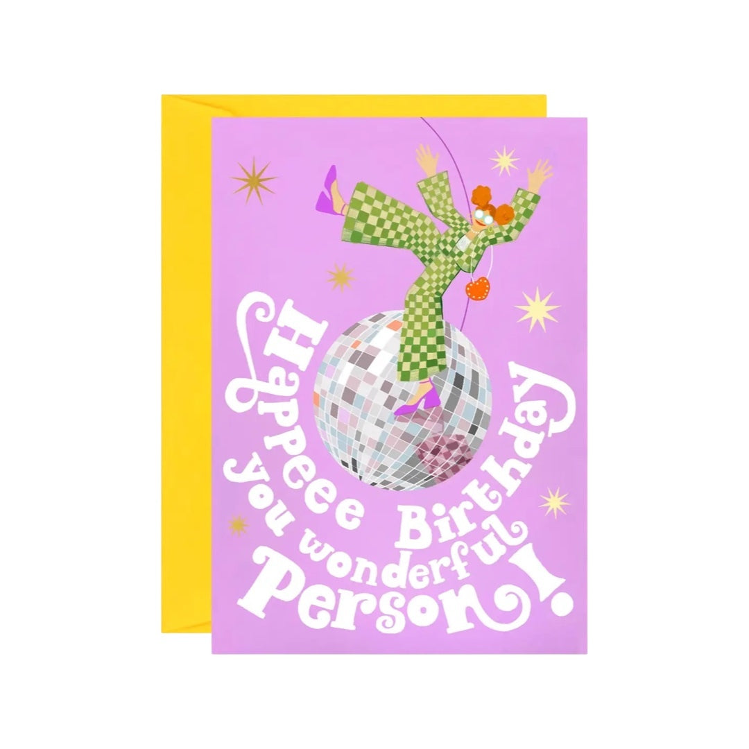 Birthday card featuring an illustration of an orange-haired lady wearing a fabulous green and white checked jumpsuit dancing on a giant glitterball and the text 'Happy Birthday You Wonderful Person' in white, all on a purple background.