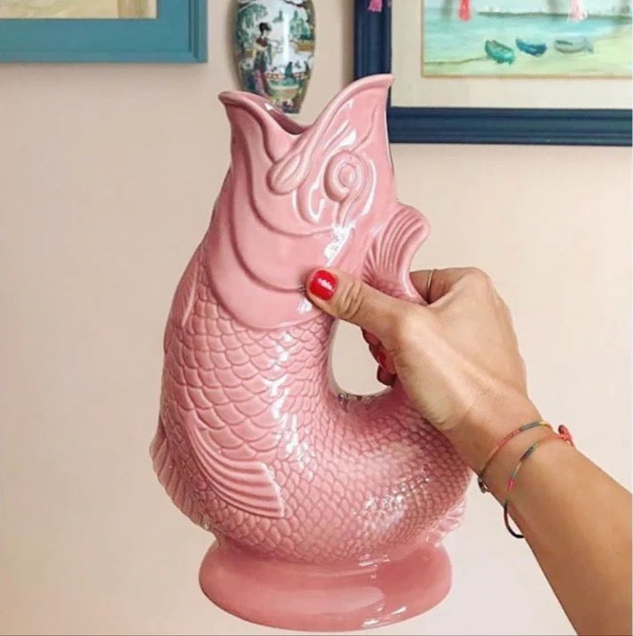 Pink fish-shaped vase held by a hand against a light-colored wall with framed pictures.