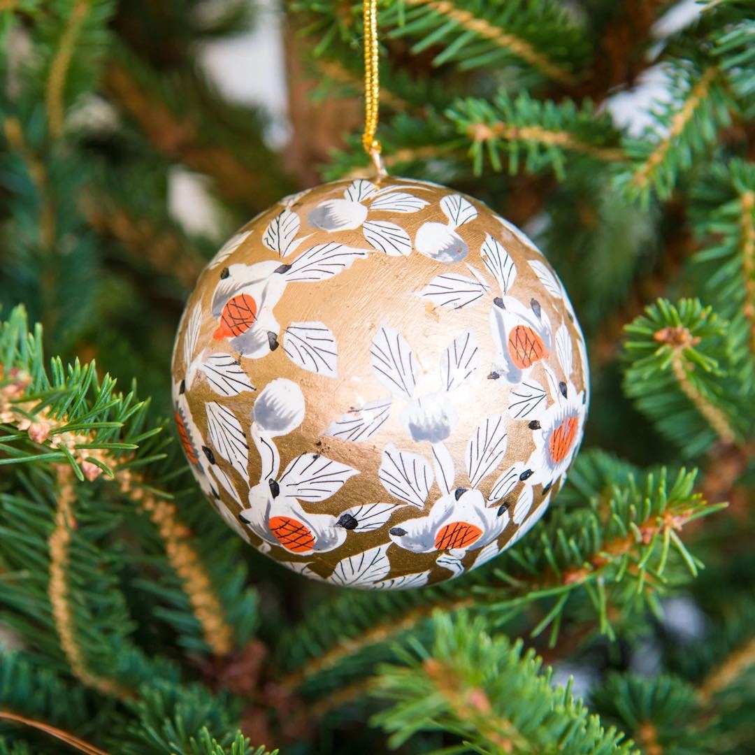 A round papier mache bauble in gold with a white, black and orange floral design hanging on a Christmas tree.