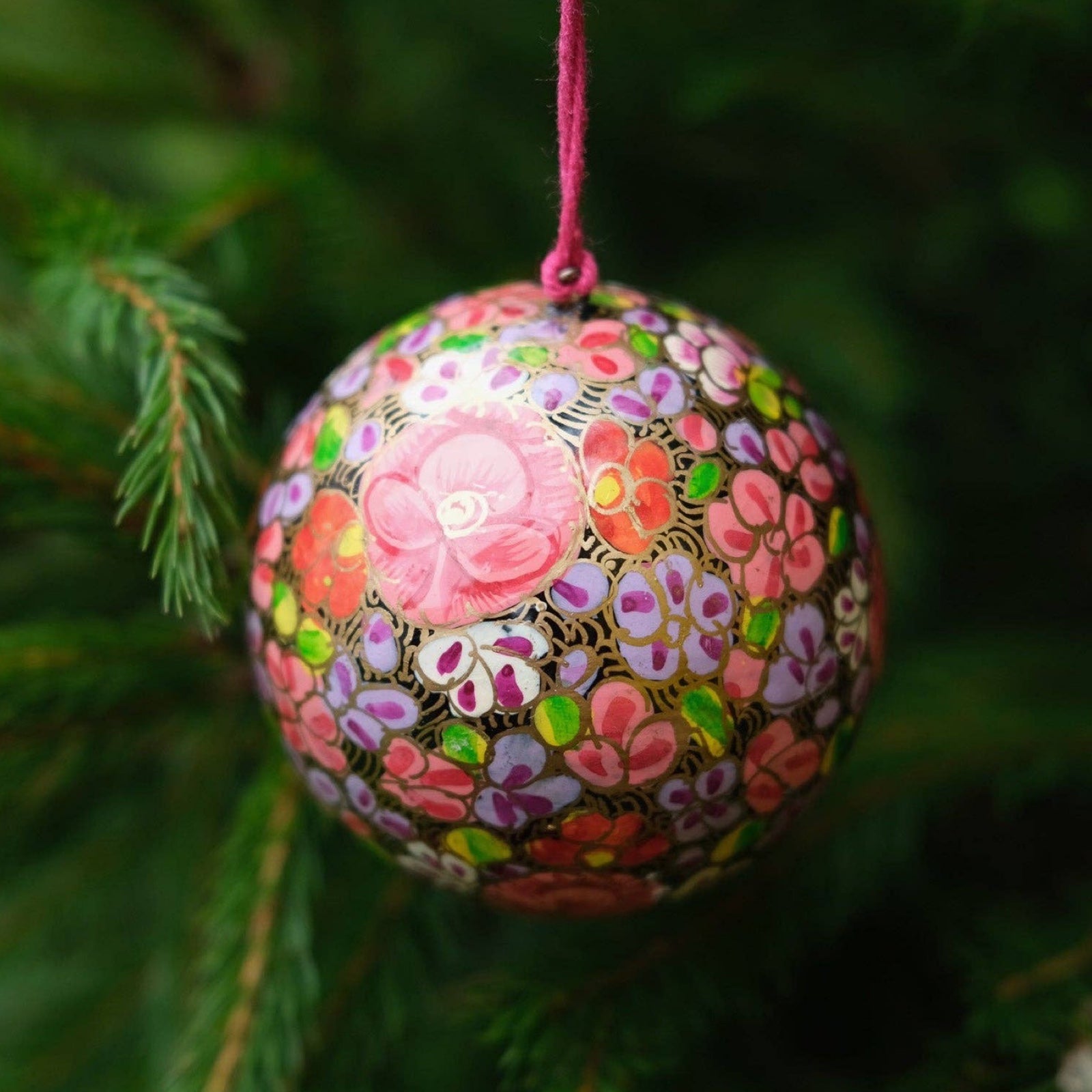 A round papier mache bauble in black with a lilac, pink, white and red floral design hanging on a Christmas tree.