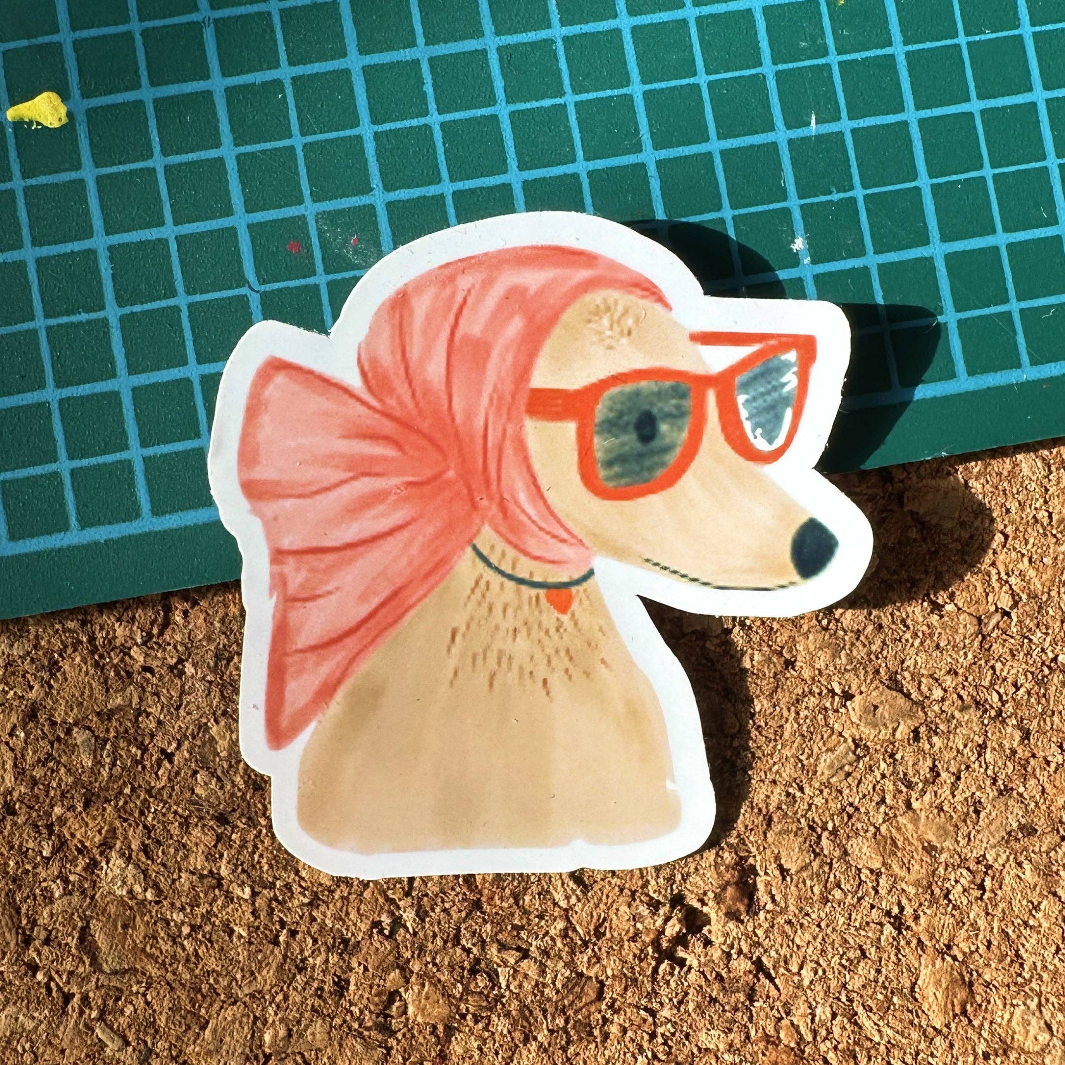 Sticker of a dog with a pink scarf and orange glasses on a cork surface.