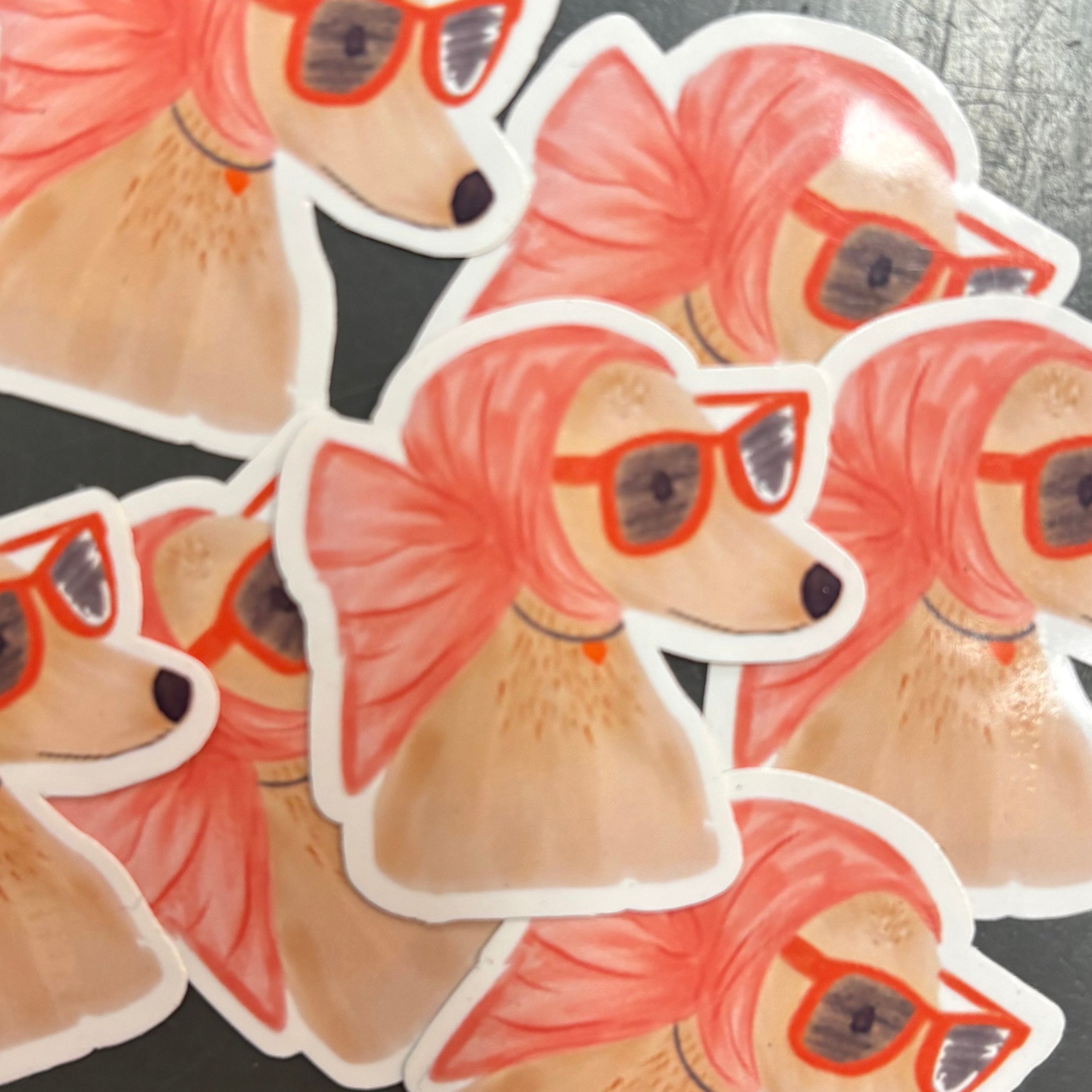Stickers of a dog with a pink scarf and sunglasses in a pile on a dark background.