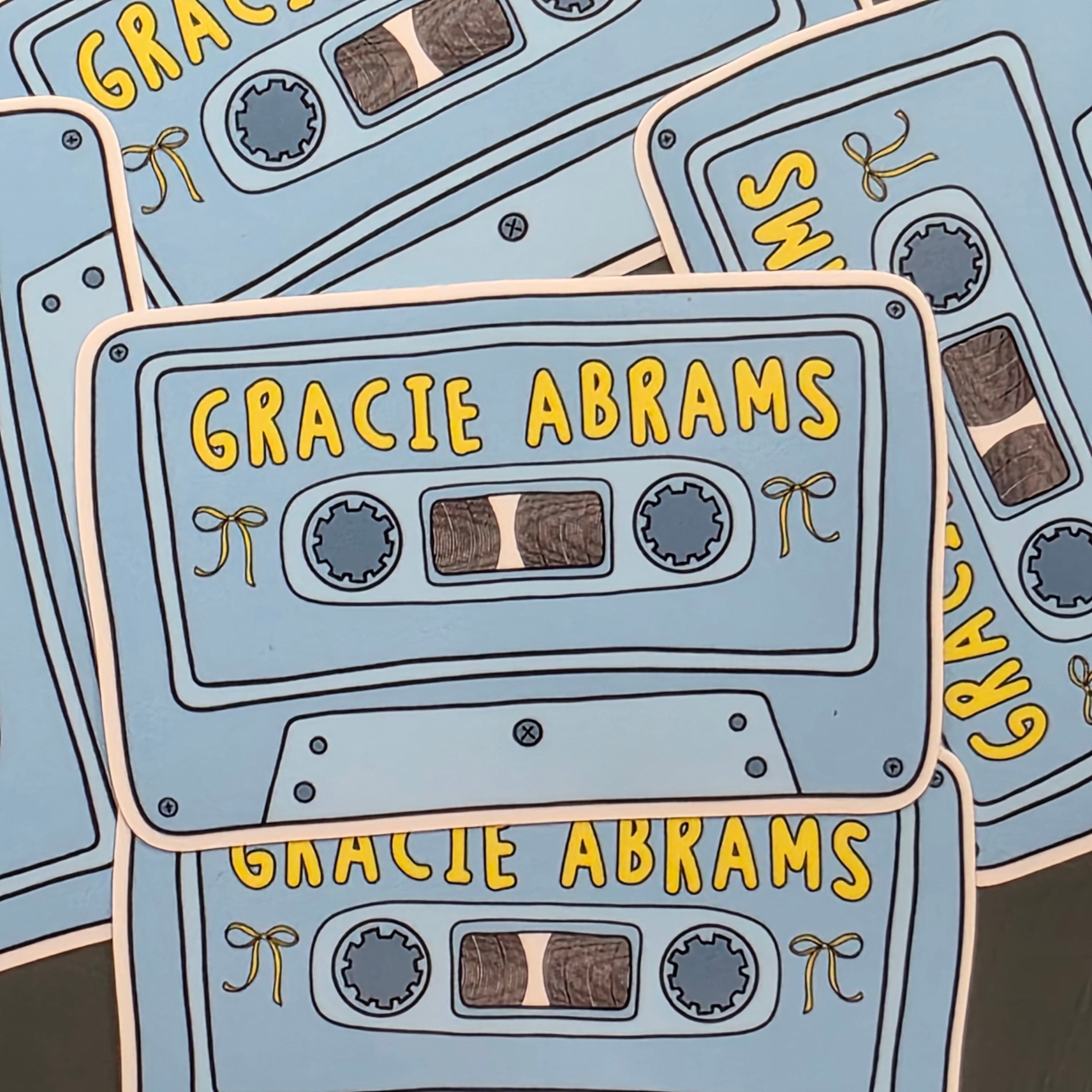 A rectangular-shaped sticker featuring an illustration of a retro cassette tape in shades of blue with the title 'Gracie Abrams', decorated with yellow bows. 
