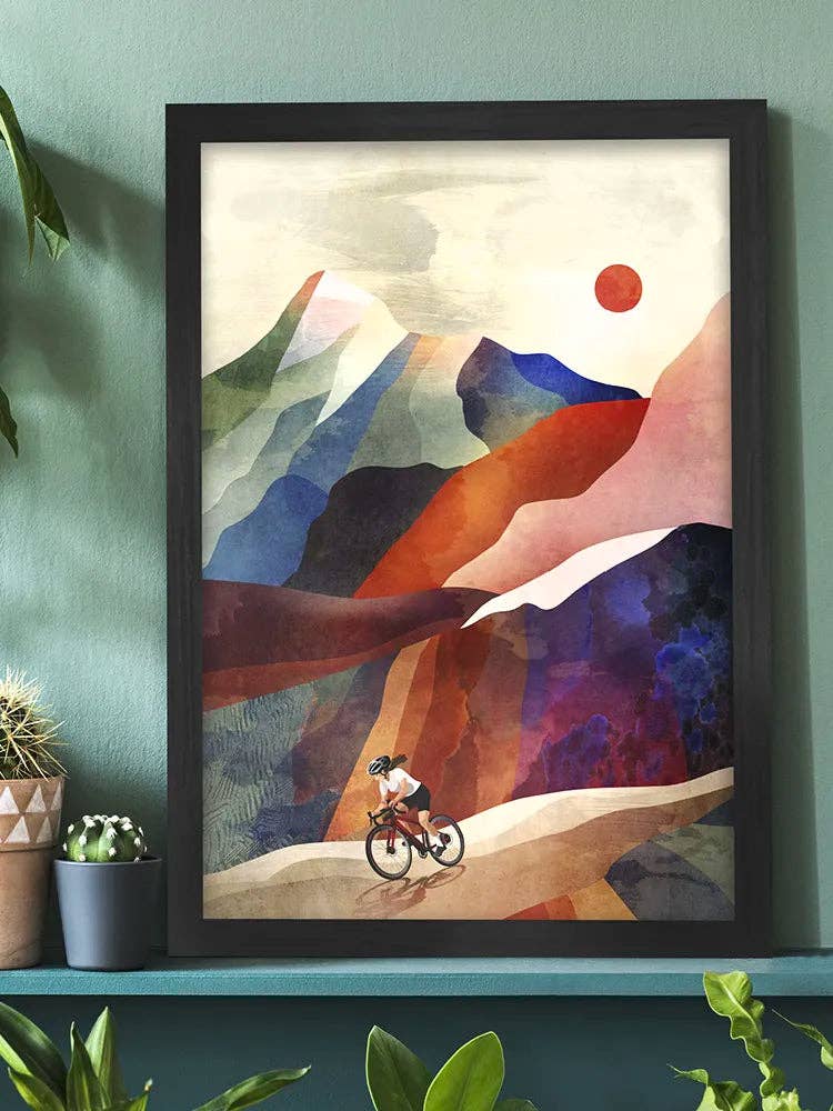 A framed abstract landscape art print featuring a cyclist on a winding path, displayed on a teal shelf against a teal wall with potted plants.