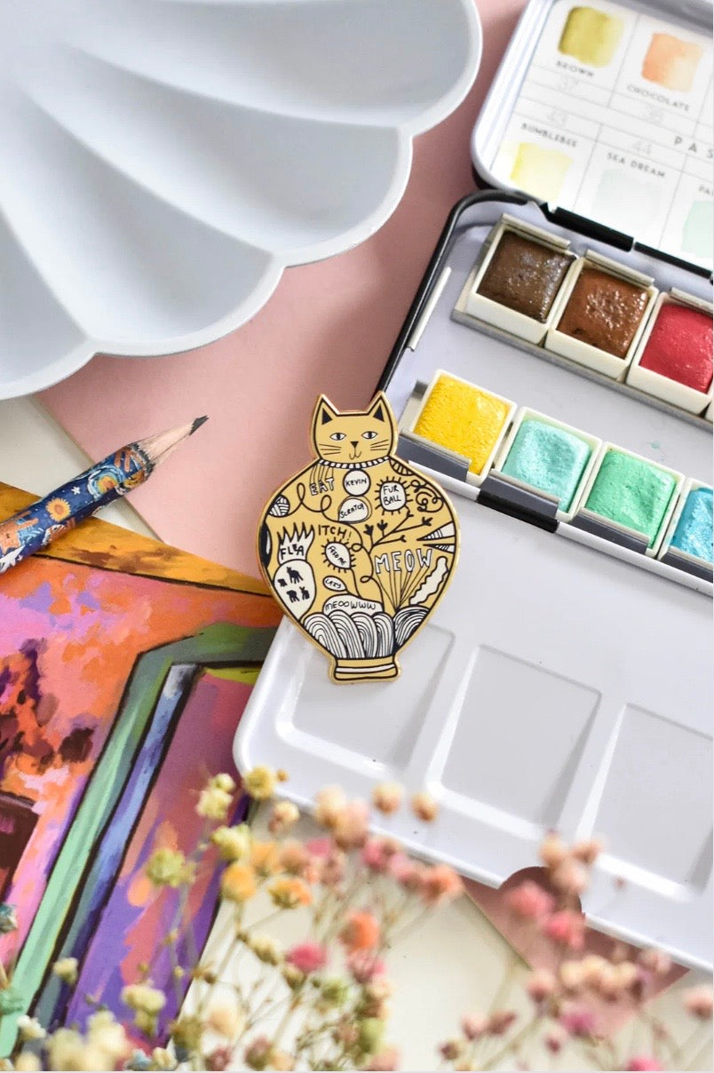 Gold cat-shaped brooch on a colourful background with art supplies.