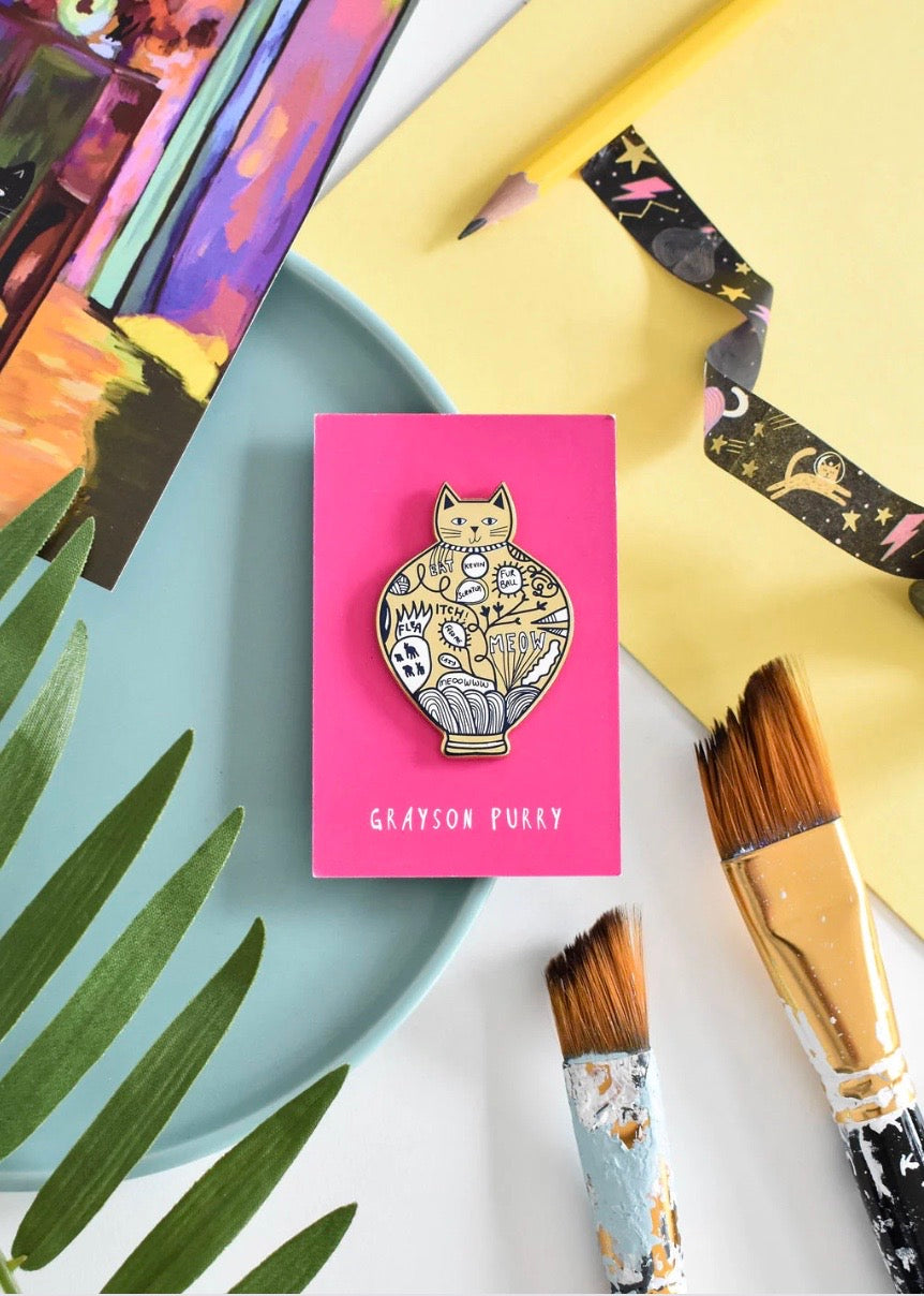 Cat-themed decorative badge on a pink card with 'Grayson Purry' branding, surrounded by artistic tools and materials.