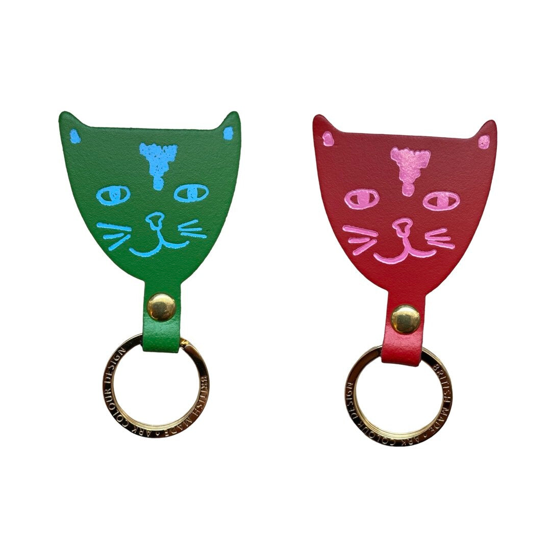 A green leather cat head shaped keyring with turquoise facial details and a red leather cat head shaped keyring with pink facial details.