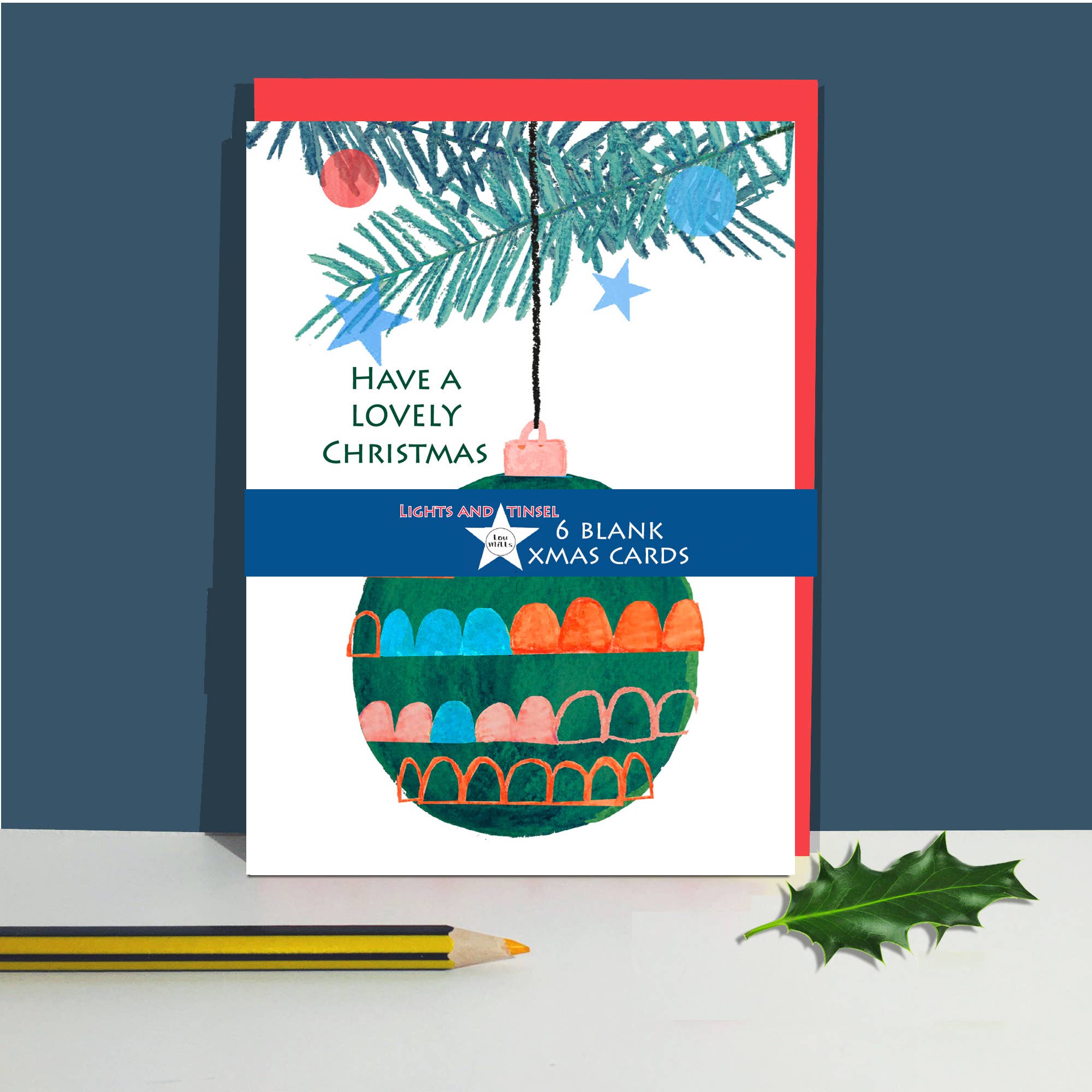 A Christmas greeting card standing upright on a white surface, with a red border, pine branches at the top, the text 'HAVE A LOVELY CHRISTMAS,' and a colourful illustrated ornament. A blue band across the middle reads '6 BLANK XMAS CARDS'.