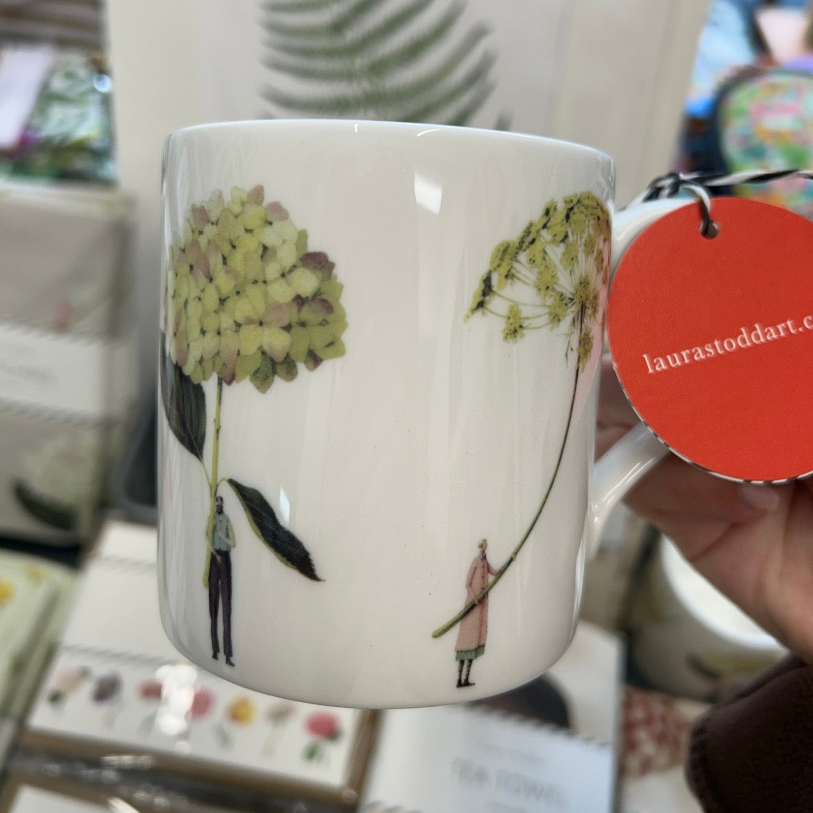 Mug with green floral design held by a hand, with a visible brand tag.