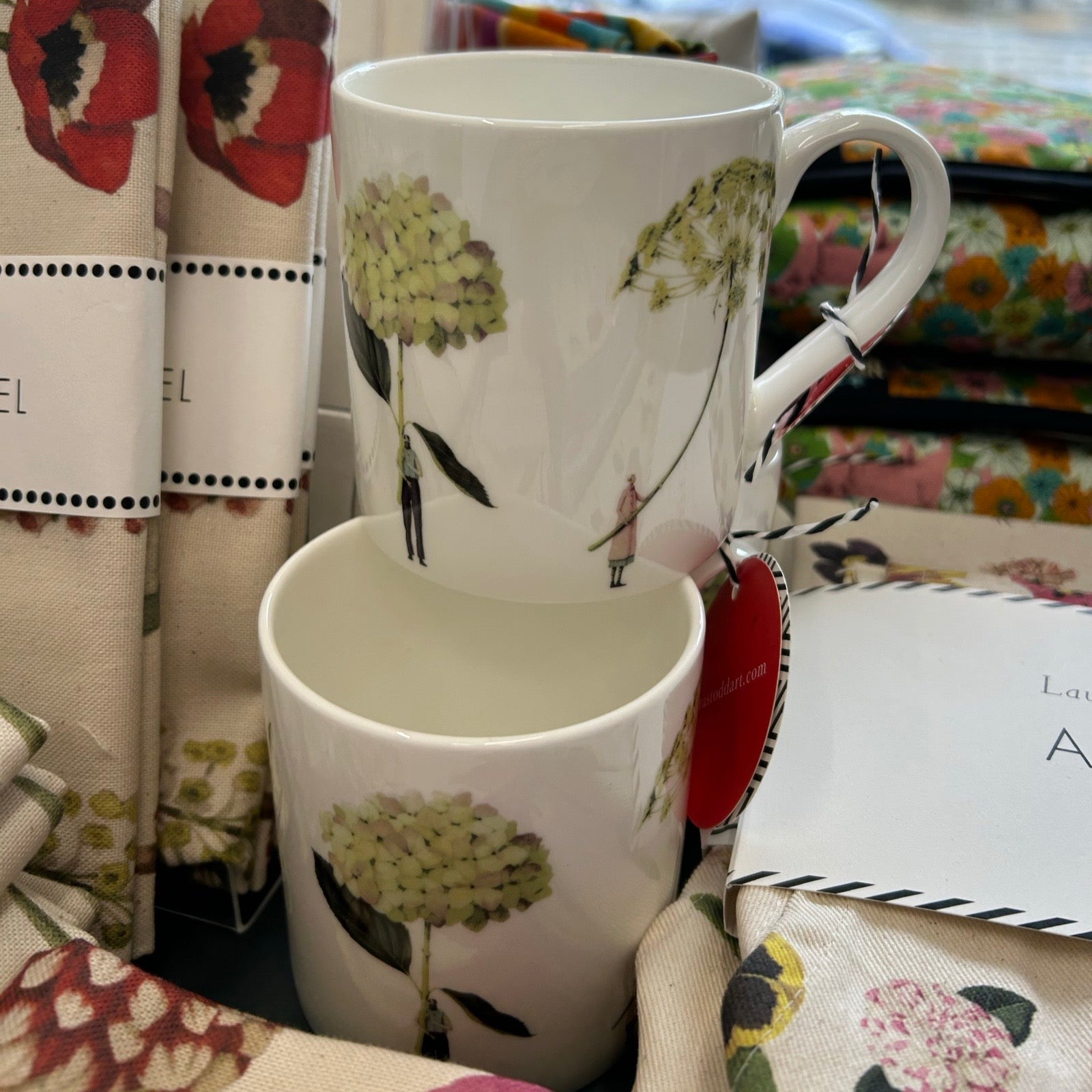 Two white mugs with floral designs on a shelf next to floral tea towels.