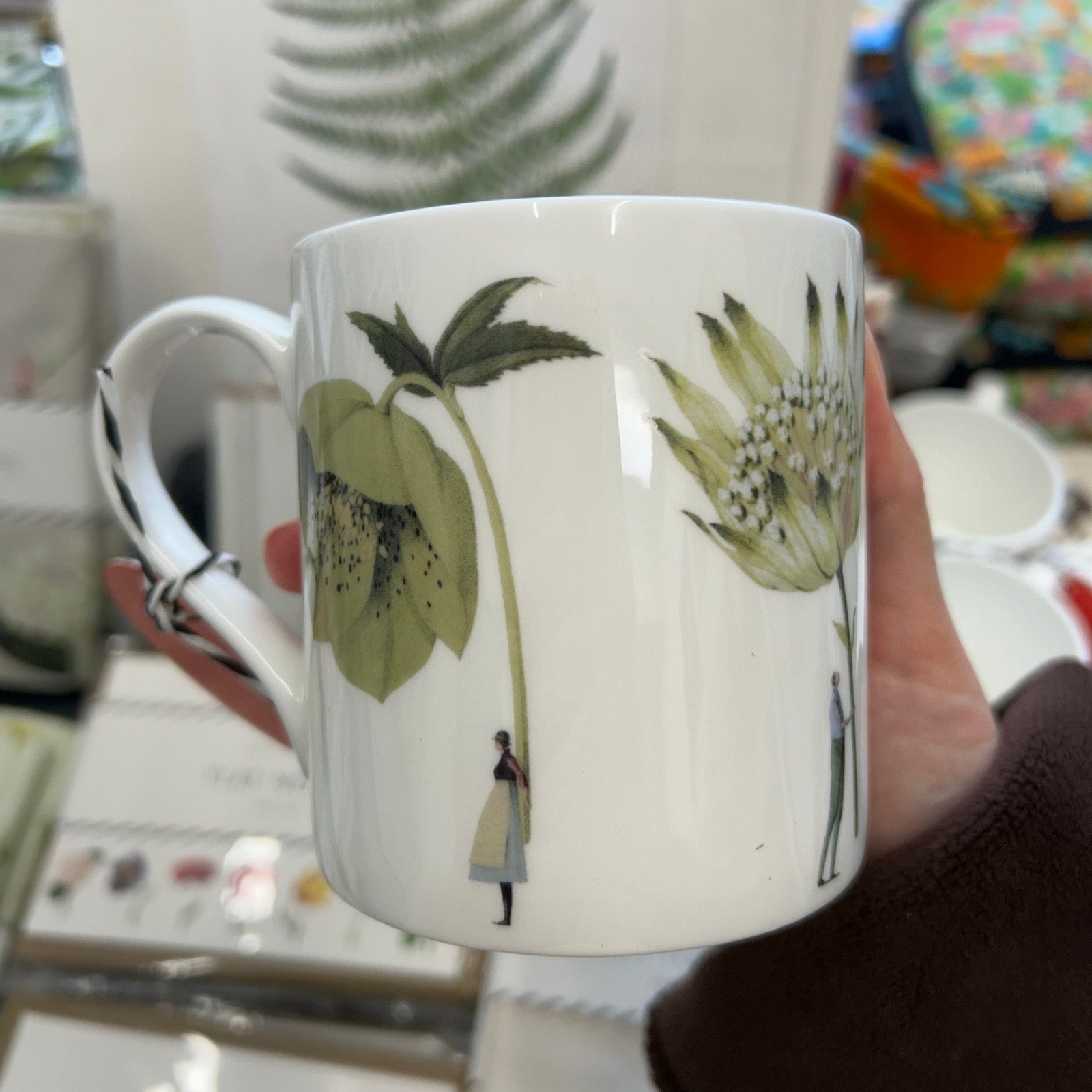 White mug with green floral design held by a hand in a store setting.
