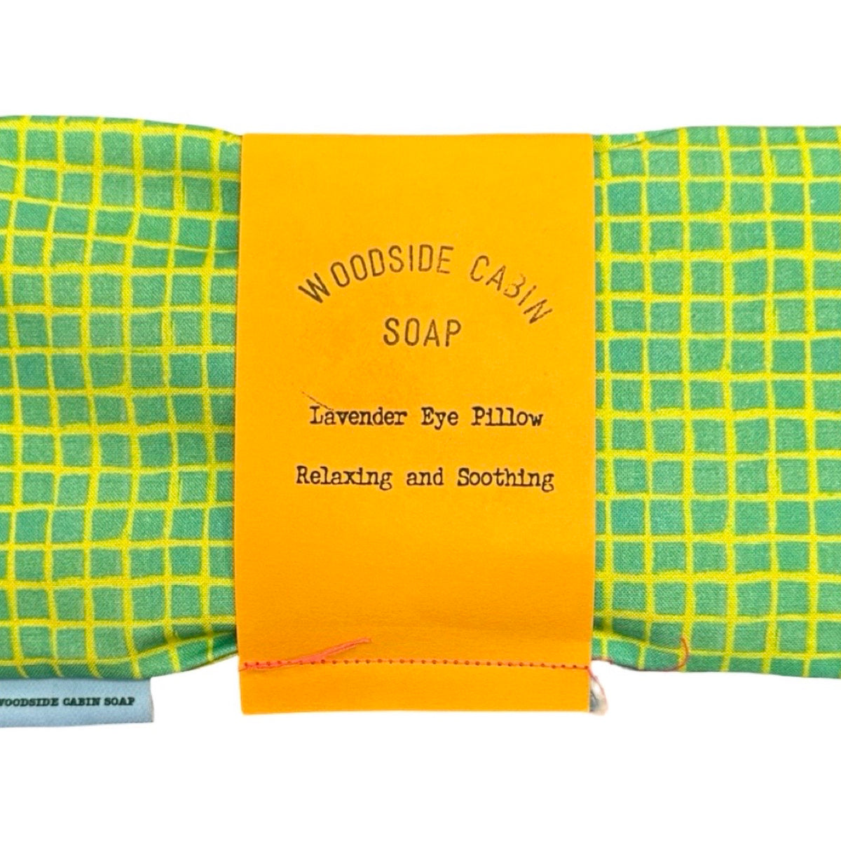 Woodside Cabin Soap lavender eye pillow in a green and yellow checkered fabric, wrapped with a bright orange paper band labeled 'WOODSIDE CABIN SOAP Lavender Eye Pillow Relaxing and Soothing'.