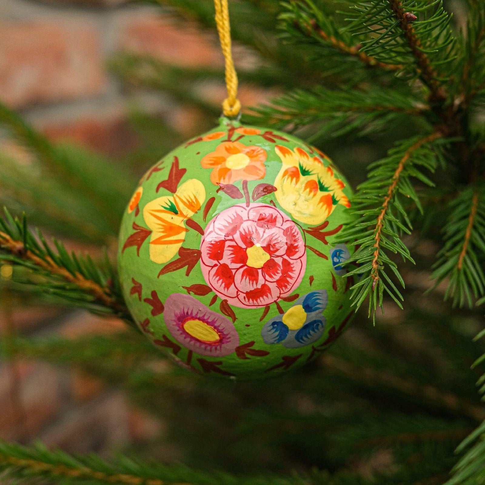 A round papier mache bauble in green with a pink, yellow, orange and blue floral design hanging on a Christmas tree.