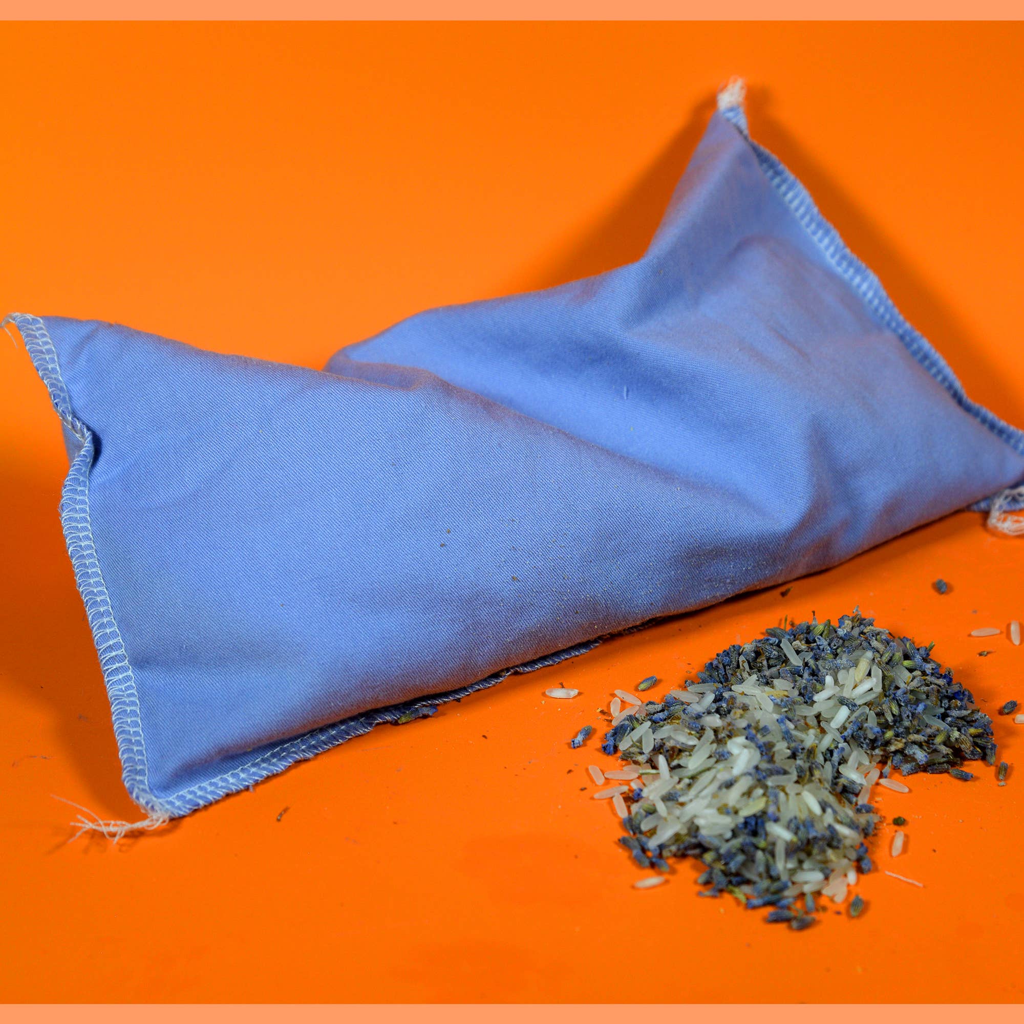A light blue fabric sachet bag with white stitching lies on an orange background, with a small pile of dried lavender buds spilled in front of it.