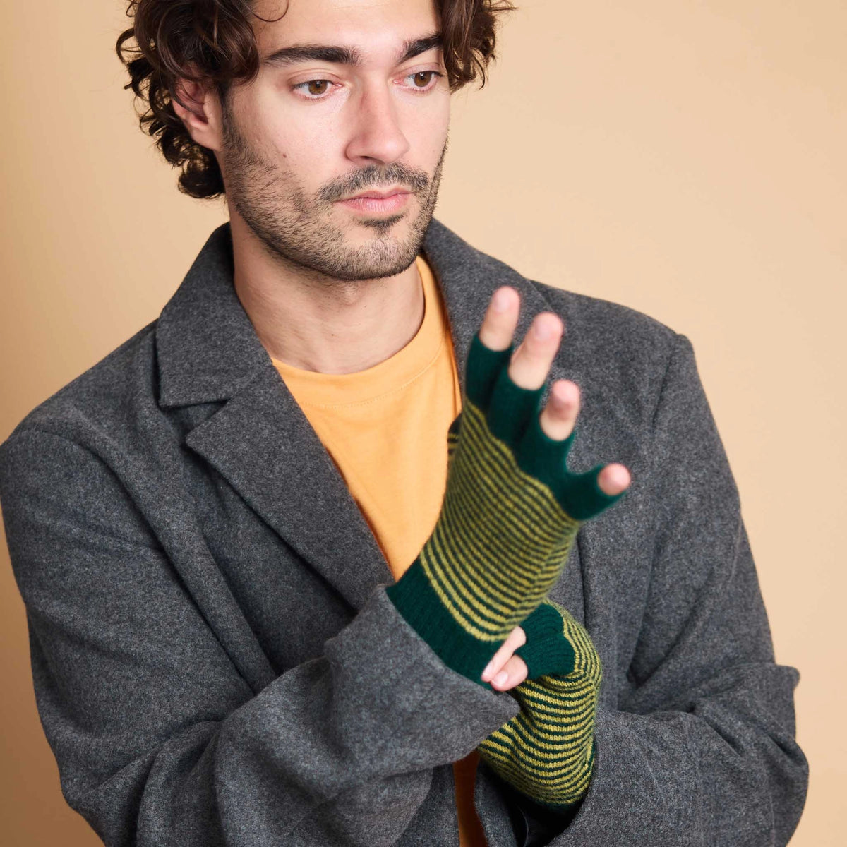 A man wearing a dark gray wool coat over a mustard yellow t-shirt, demonstrating a green and yellow striped knit fingerless glove on his hand against a solid beige background.