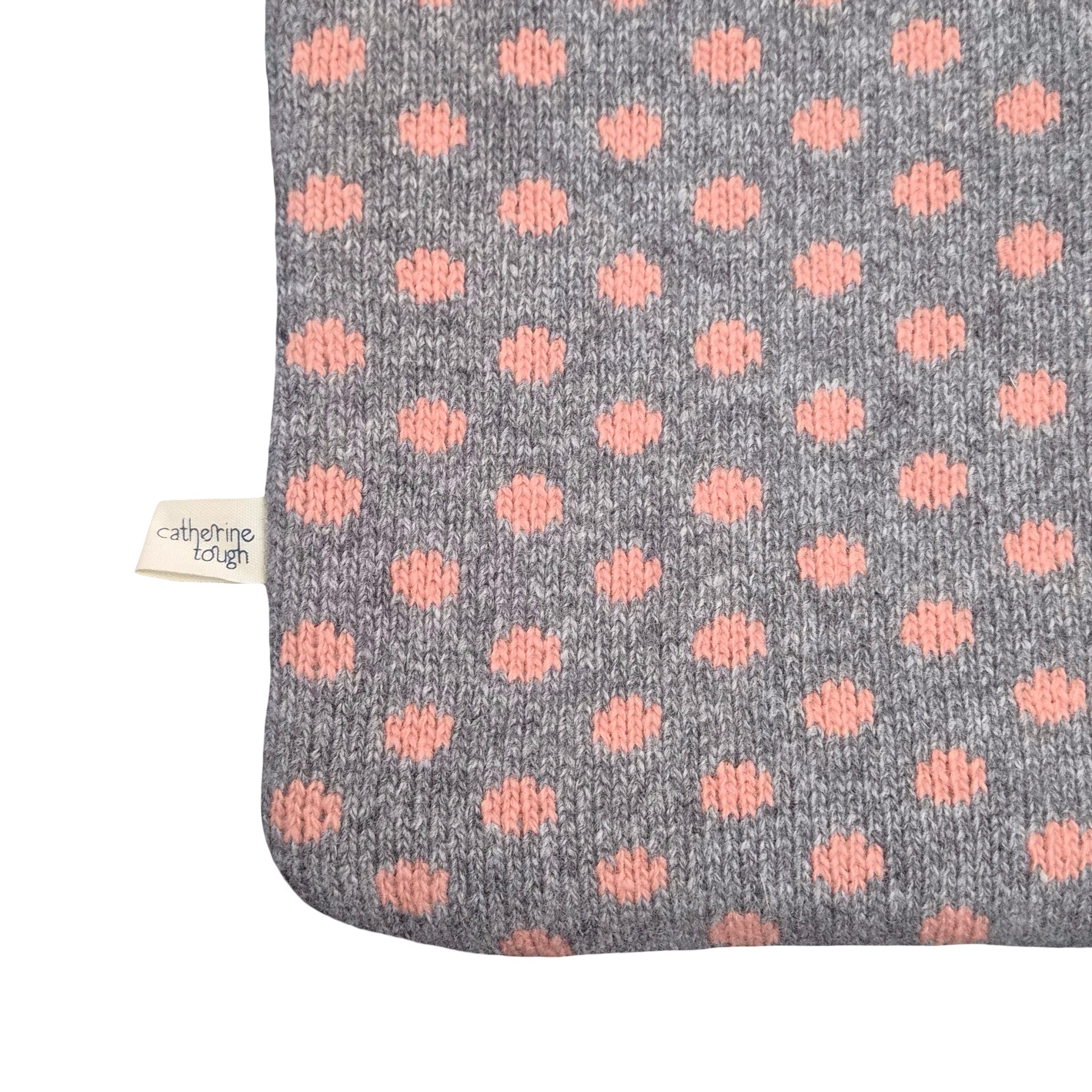 A close-up of a knitted hot water bottle in grey with a repeating pattern of dusty pink polka dots, featuring a small white fabric tag on the left side.