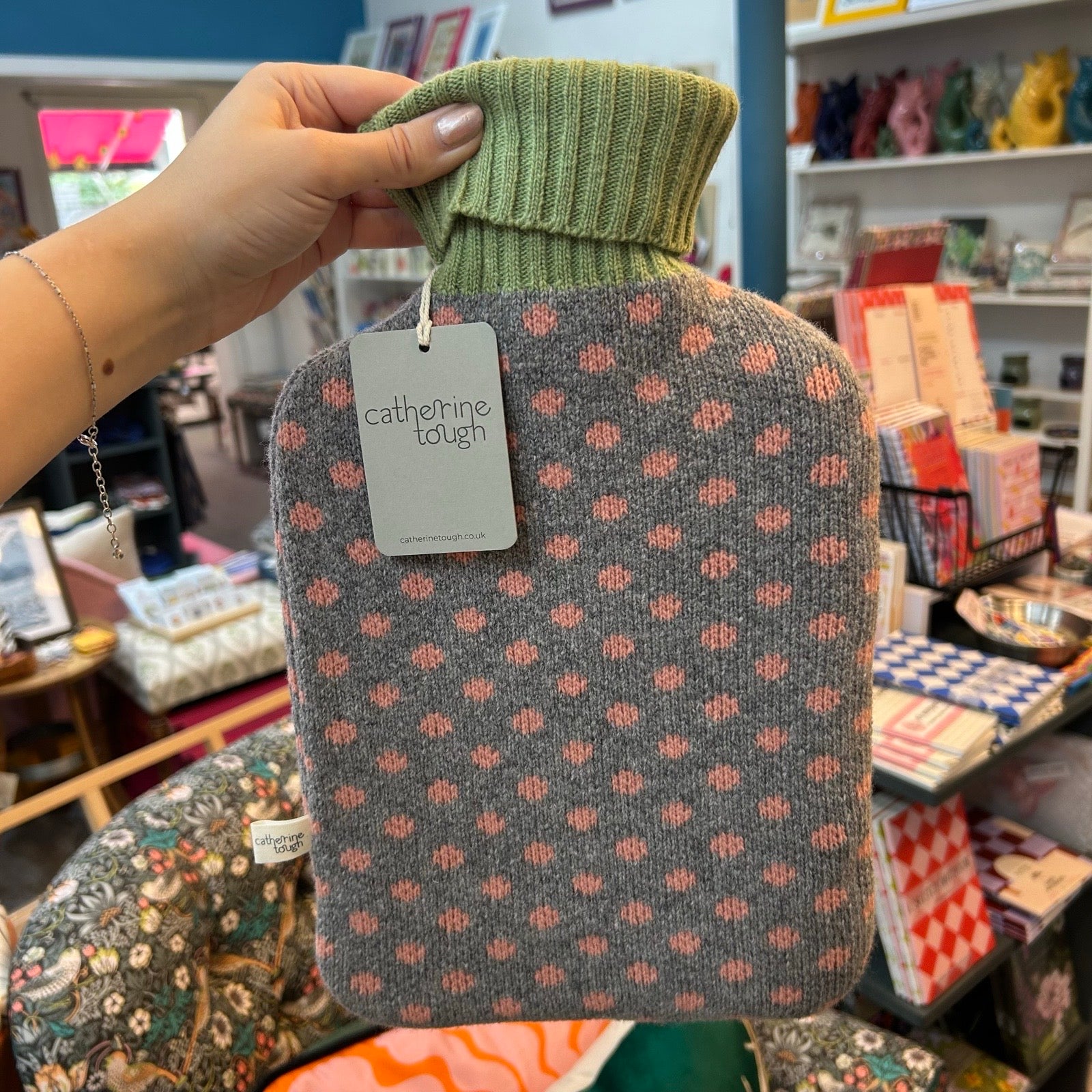 A hand holds a hot water bottle covered by a grey knit sleeve with a pink spots pattern and a green ribbed turtleneck. A tag reads 'catherine tough'.