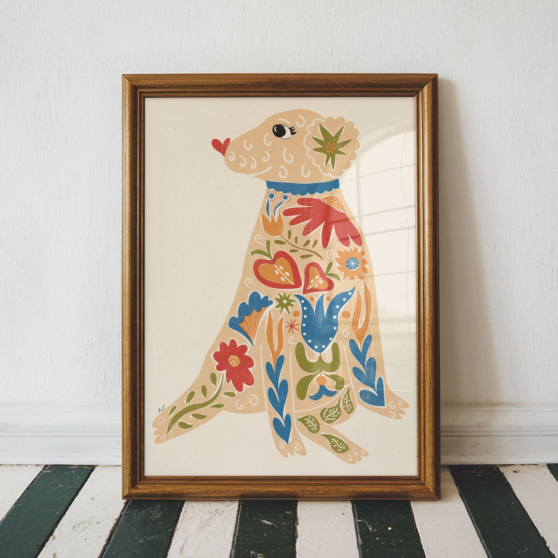 A framed folk art print of a seated dog decorated with colourful floral patterns, leaning against a white wall on a black and white striped wooden floor.