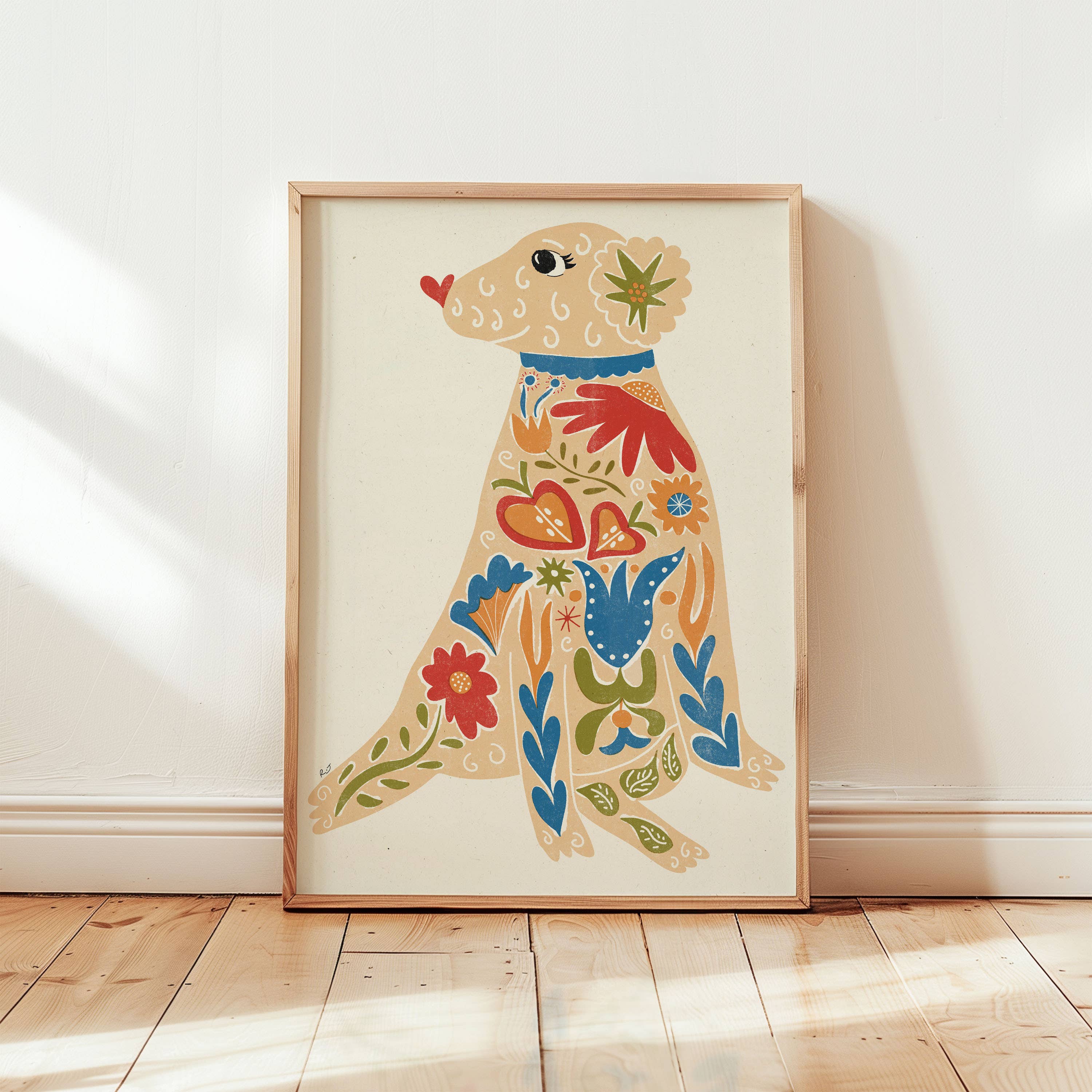 A framed folk-art print of a seated dog decorated with colourful flowers and patterns, leaning against a white wall on a light wood floor.