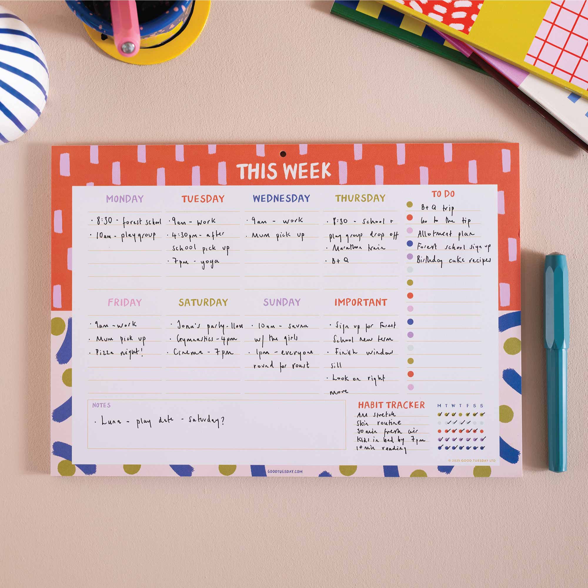 A weekly planner notepad with a colourful patterned border and a structured weekly schedule layout, photographed on a beige desk with school and office props around it.
